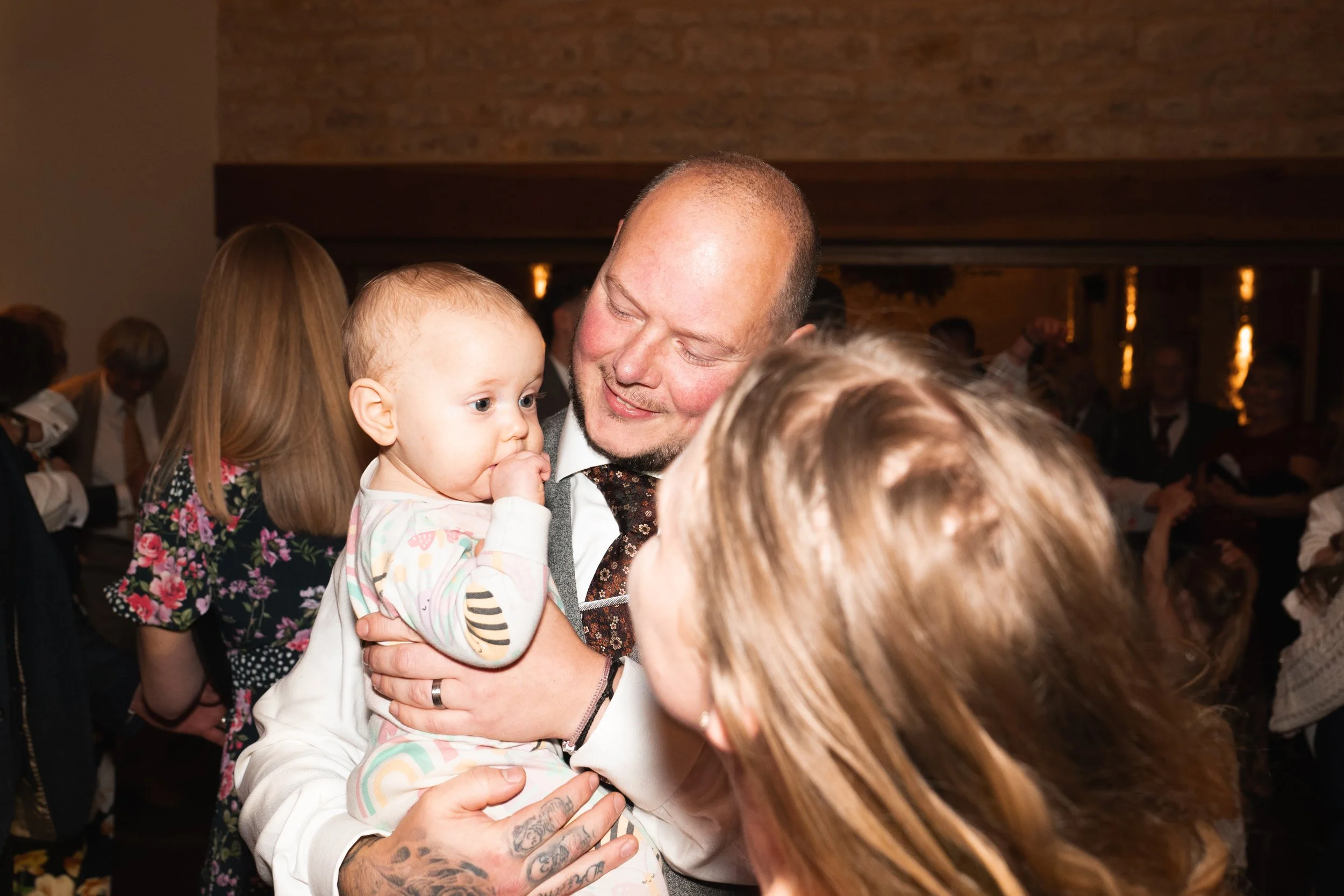 A man holding a baby girl, smiling in a crowded indoor setting with warm lighting.