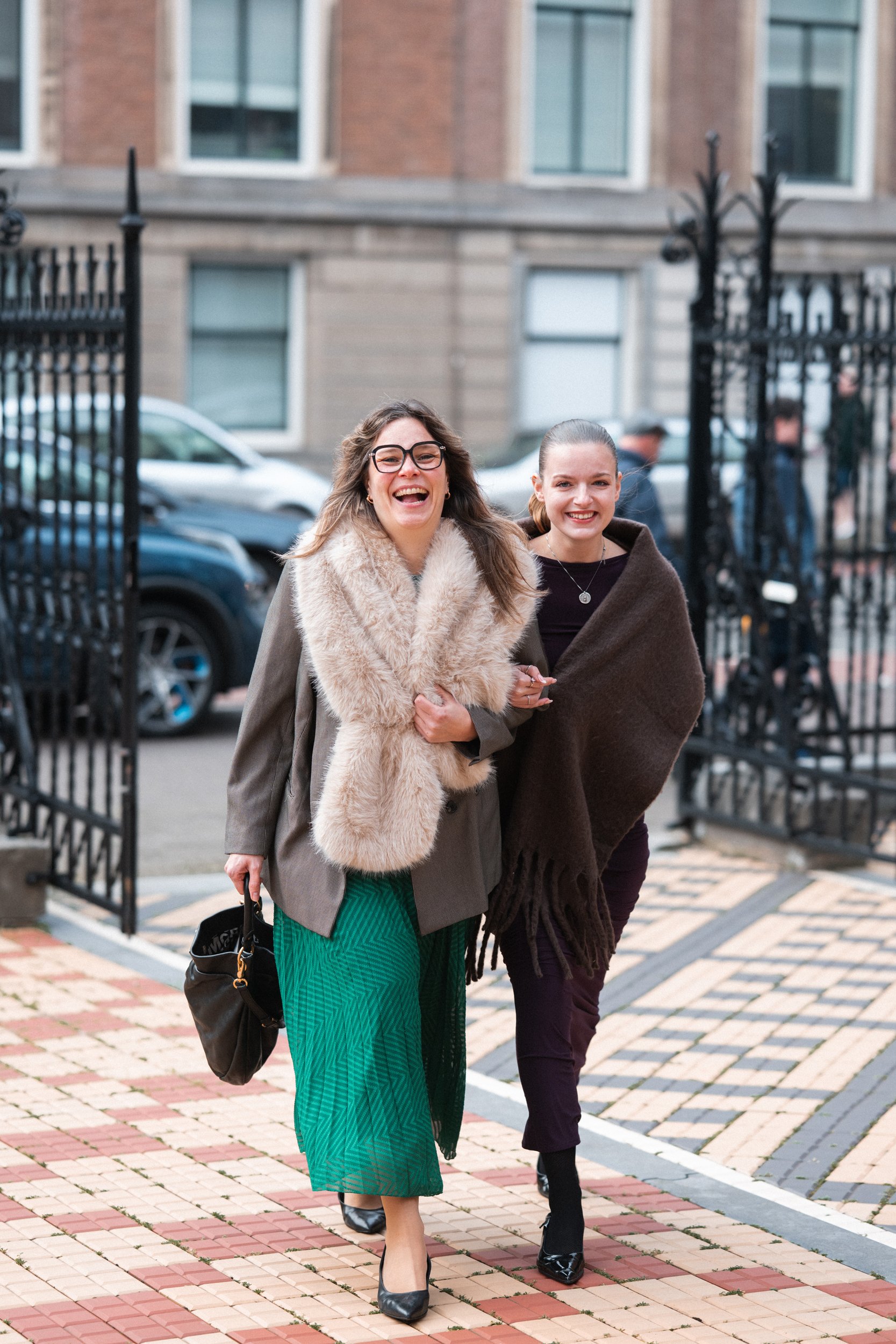 Two women walking happily outdoors, dressed in stylish winter clothing, with a city building and parked cars in the background.