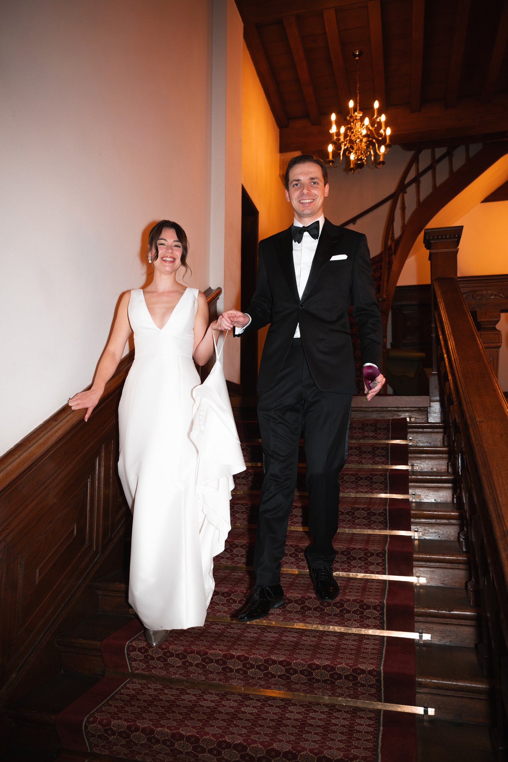 A woman and man in formal attire walking down a staircase, smiling. The woman is wearing a white gown, and the man is wearing a black tuxedo. The setting appears to be a fancy venue with wood-paneled walls and a chandelier hanging from the ceiling.