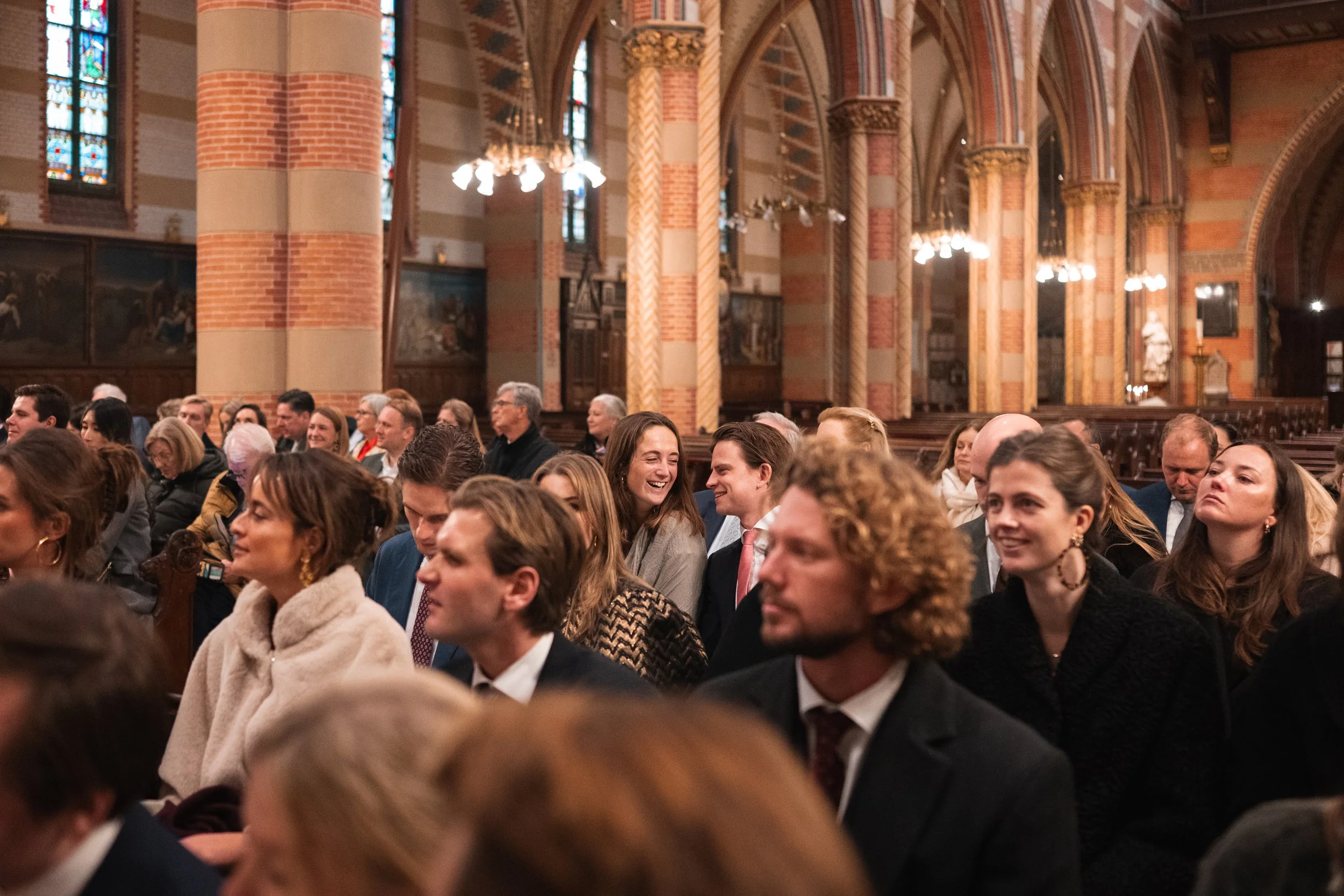 People sitting in pews inside a church, some smiling and talking, with ornate architecture, columns, stained glass windows, and chandeliers.