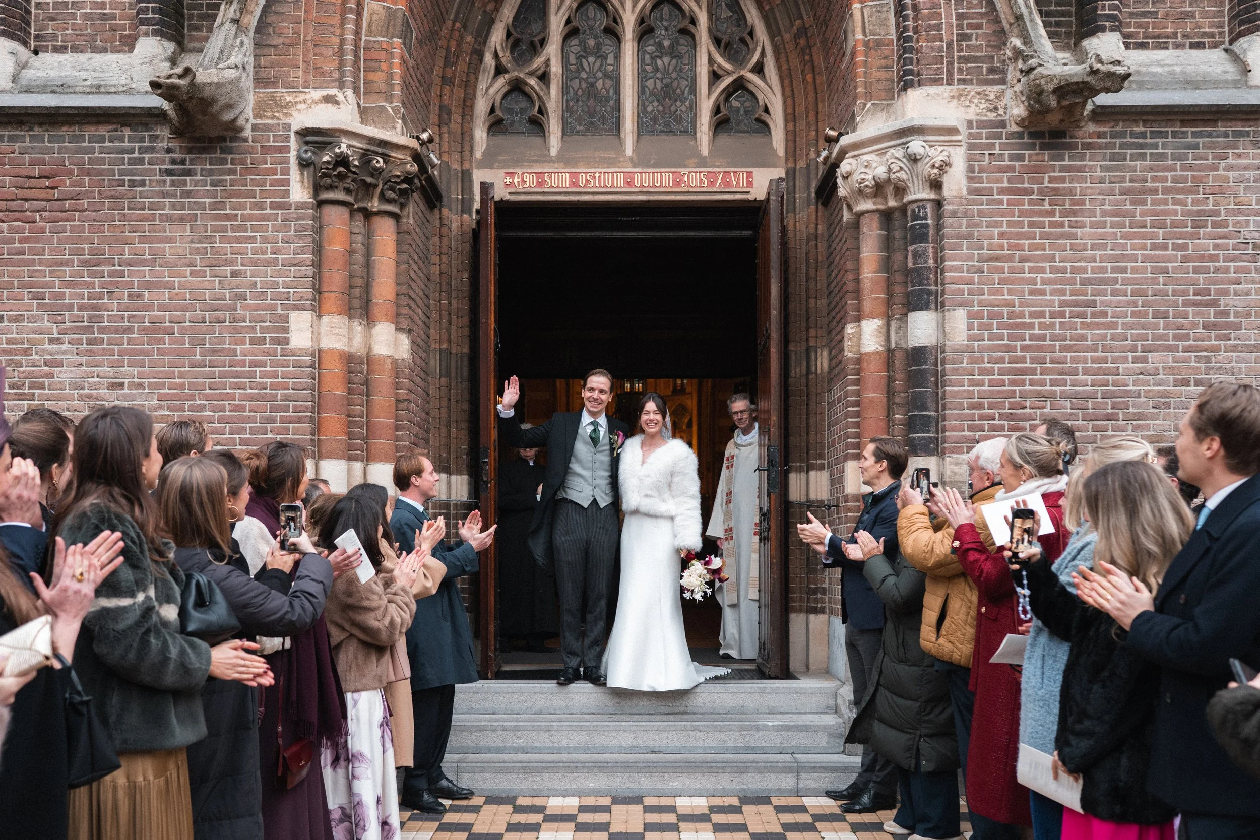 A newly married couple standing at the entrance of a church, waving to guests. The bride is wearing a white wedding dress with a fluffy white shawl and holding a bouquet, while the groom is in a dark suit with a green tie. Guests are clapping and tak