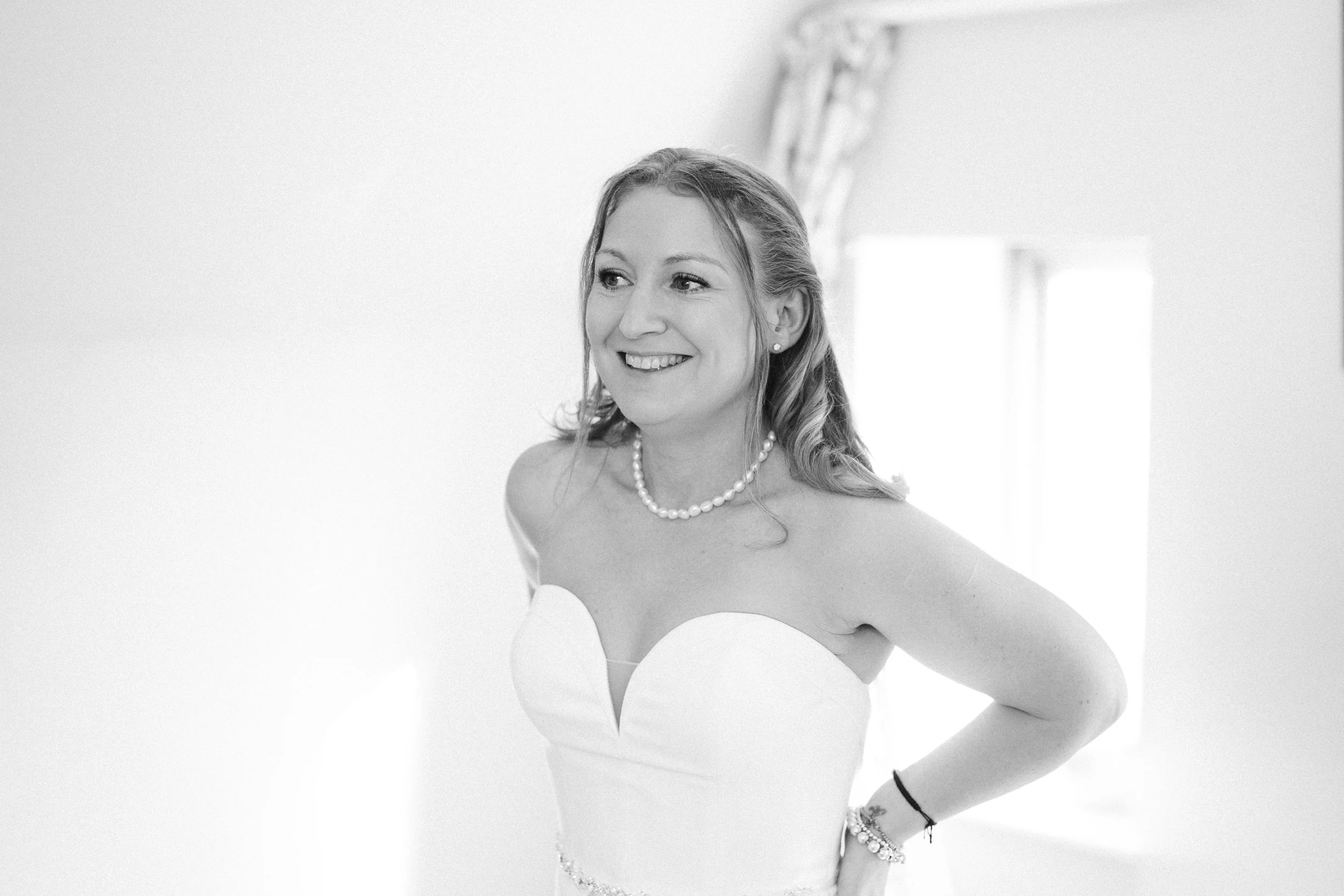 A woman in a strapless wedding dress with pearl jewelry, smiling and standing with her arm on her hip in a bright room.