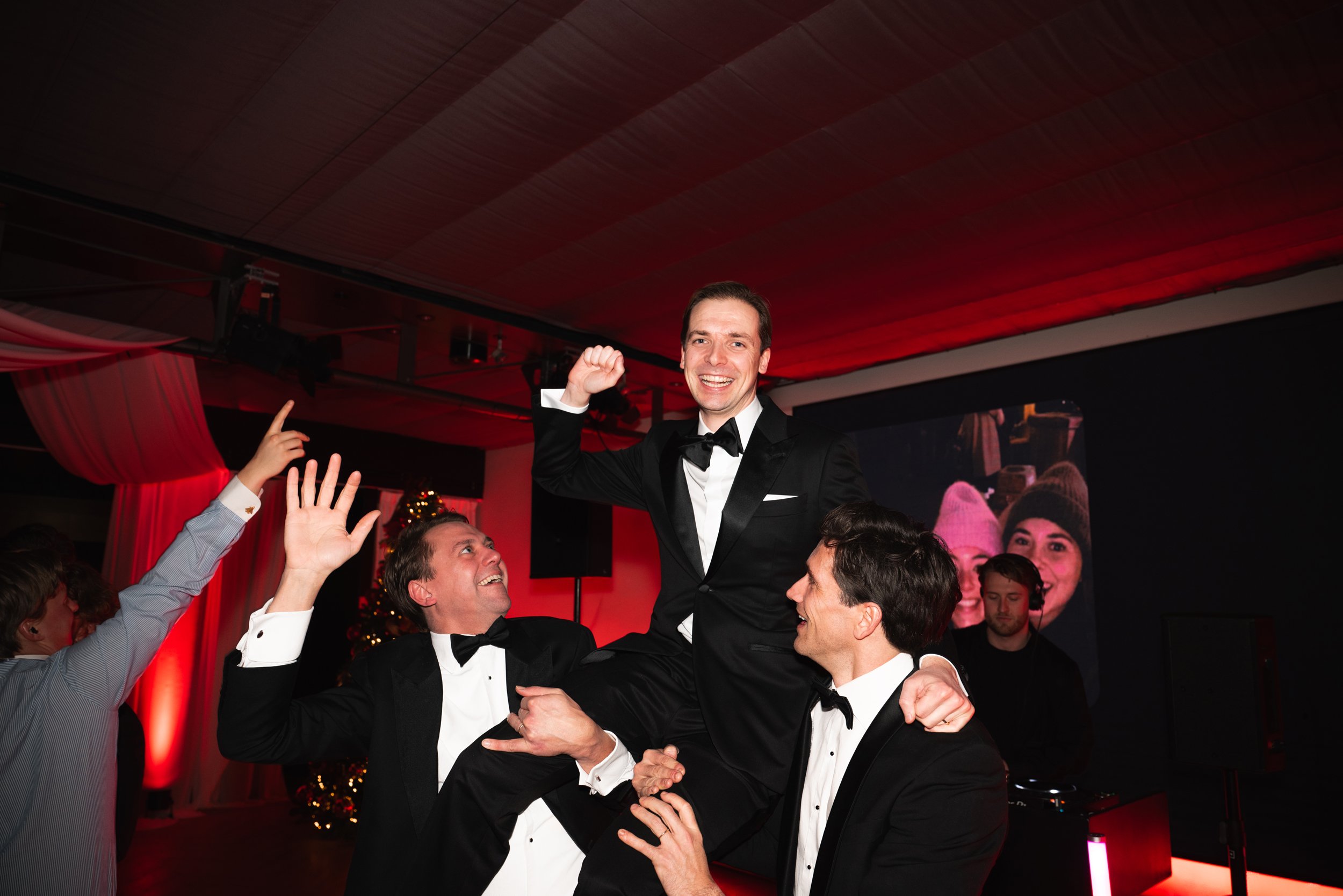 Group of men in tuxedos celebrating, with one man being lifted up and several people reaching out towards him, in a festive indoor setting with red lighting and holiday decorations.