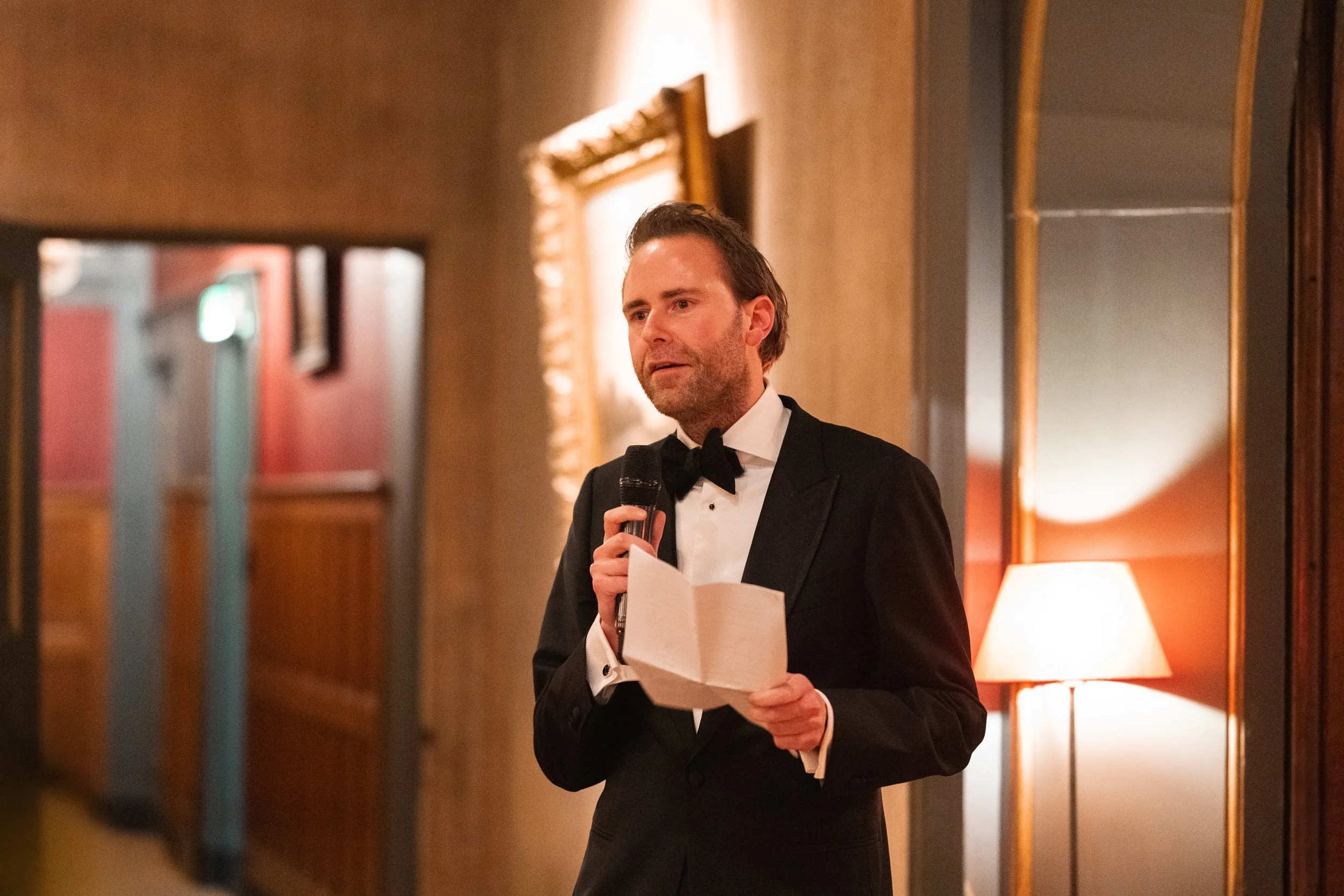 A man in a tuxedo holding a microphone and a paper, standing in a warmly lit room with wooden walls and framed art.
