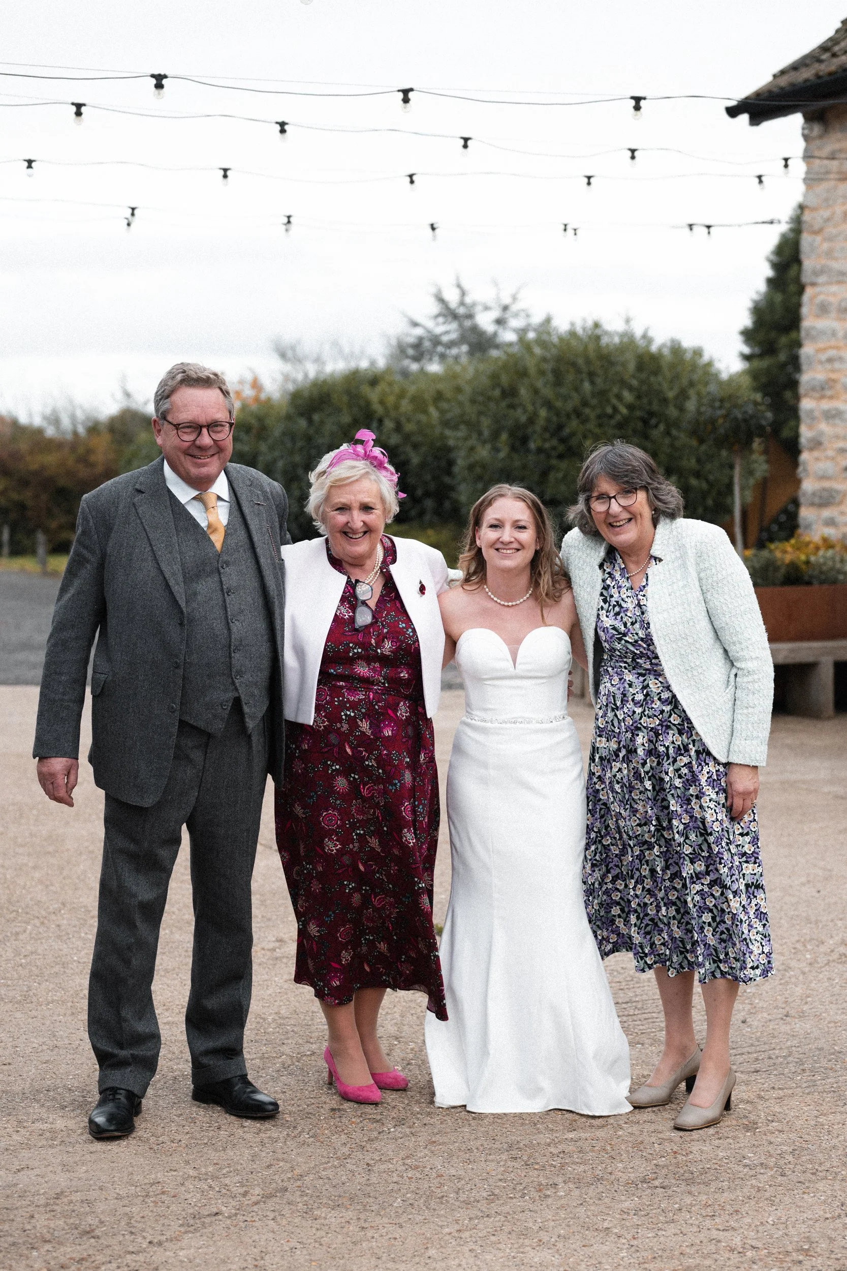 Group of four people smiling at a wedding outdoors, with a bride in a white strapless dress in the center, flanked by two women and a man, all dressed in formal attire.