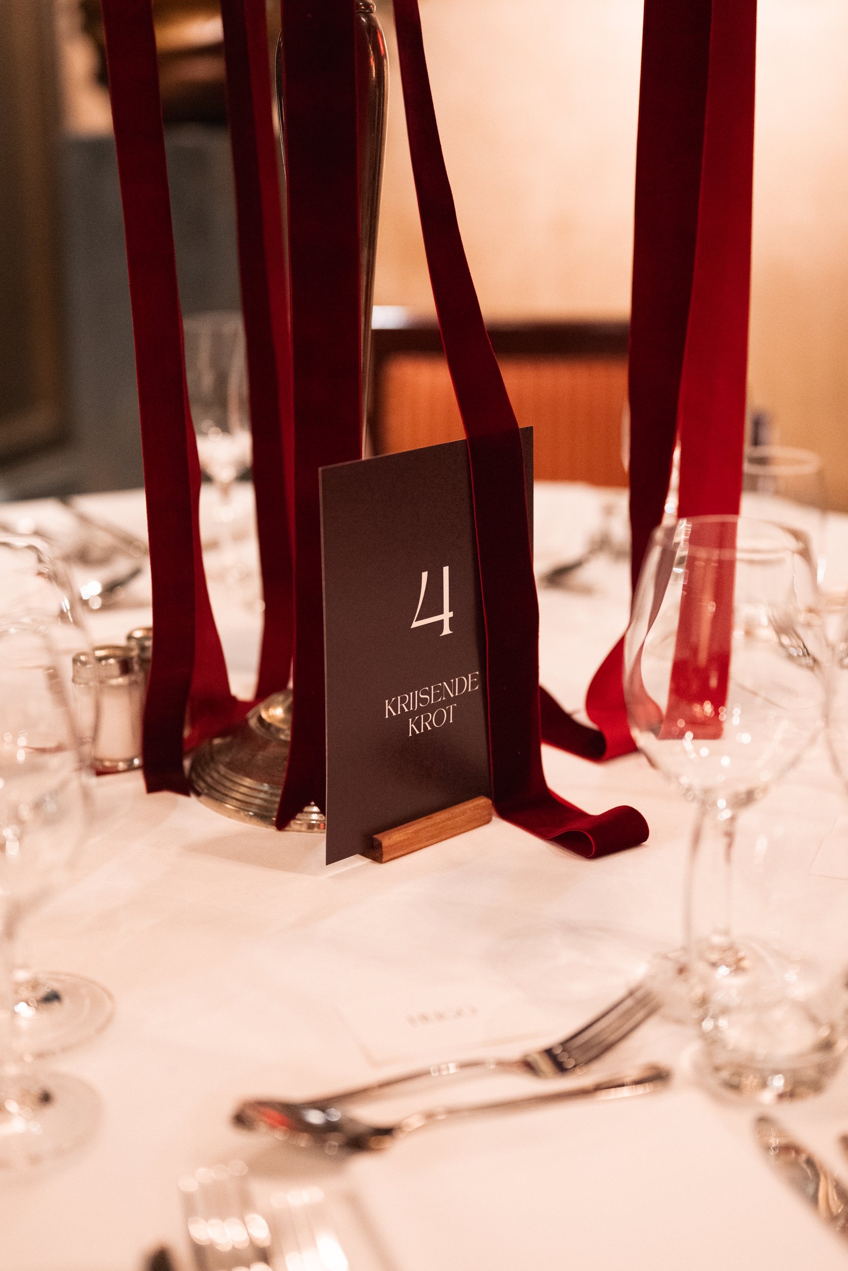 Table setting at an event with a black and white table number card displaying number 4 and the words 'Krisende Krot', surrounded by glassware, silverware, and red ribbons, with a centerpiece.