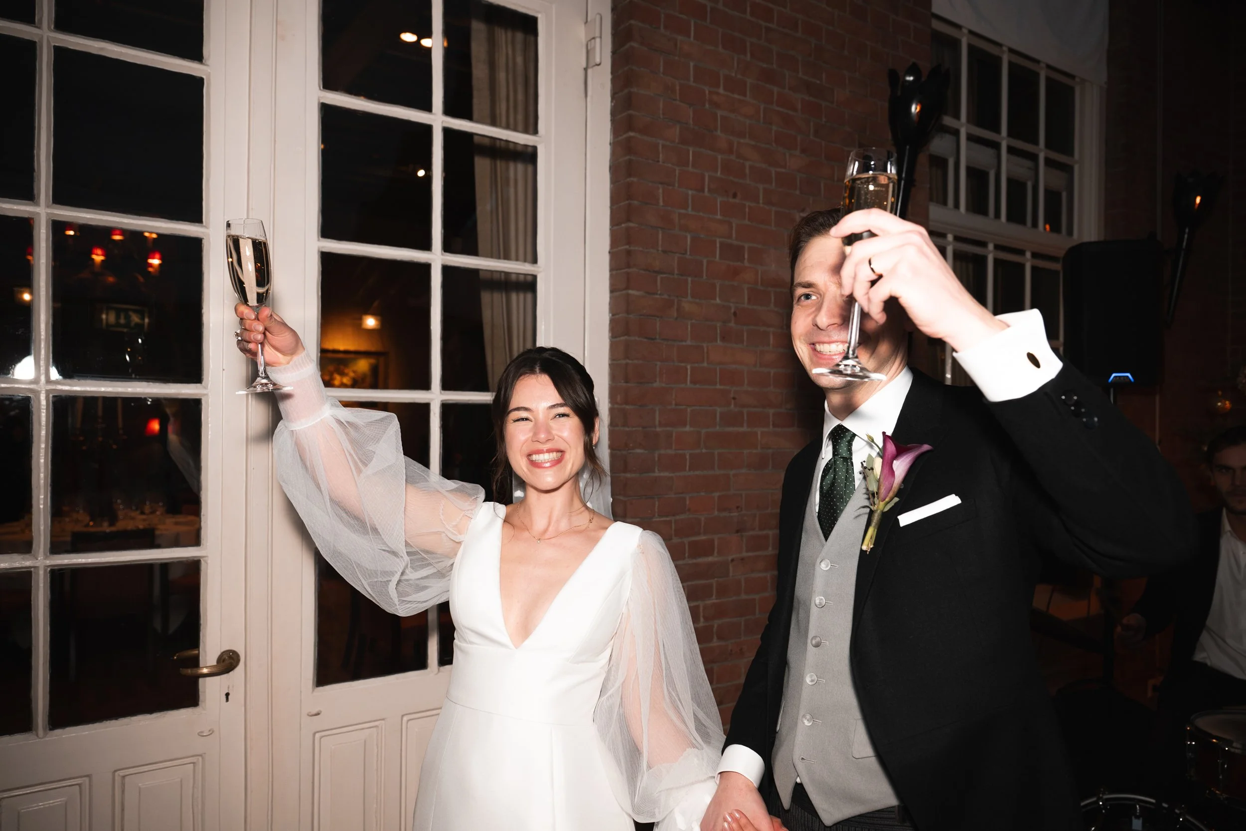 A smiling bride and groom at their wedding reception, holding champagne glasses, with the bride raising her glass and the groom holding his near his face.