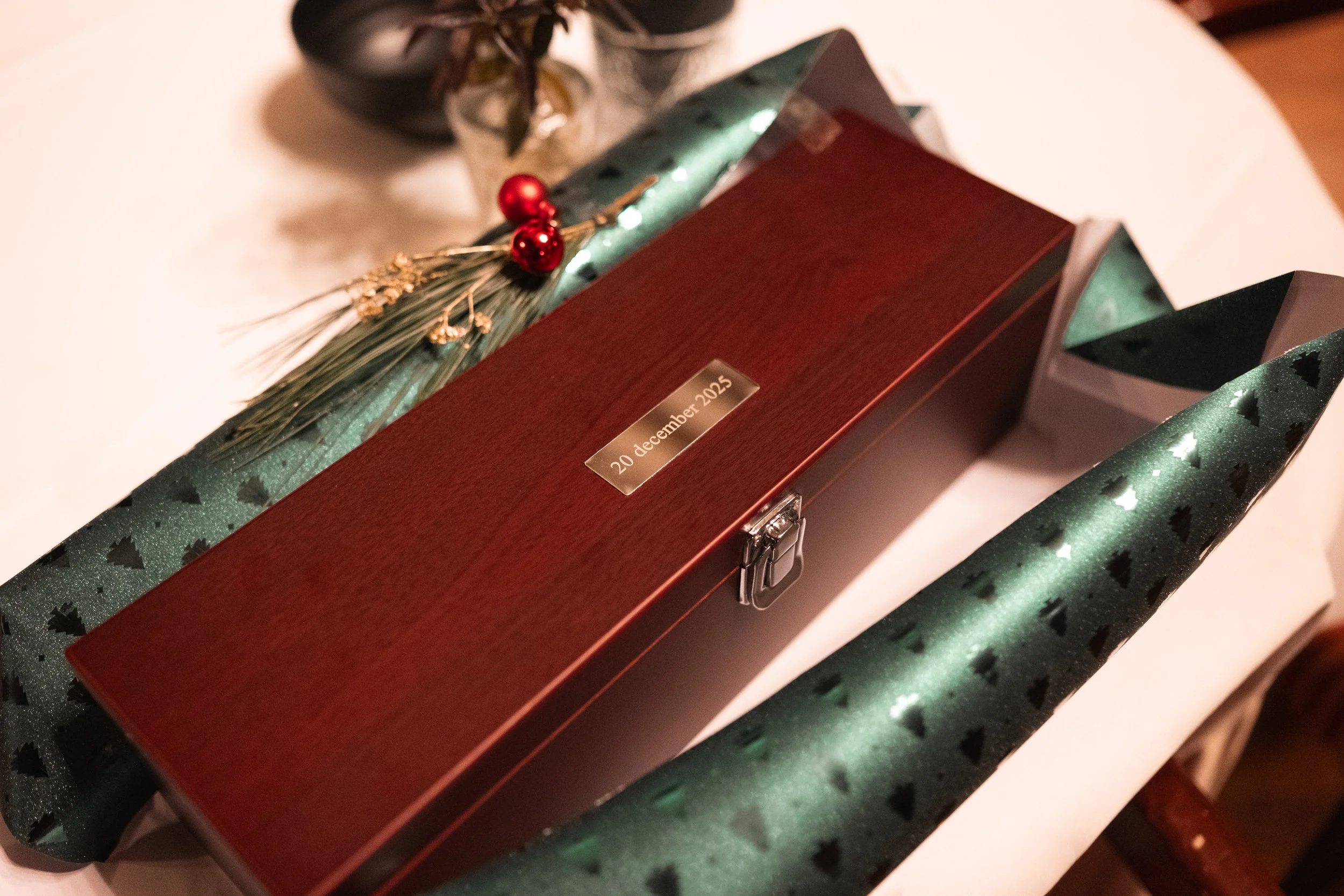 A wrapped gift box with a dark red wooden lid, a small metallic tag with the date '20 December 2025', and a lock. The gift is decorated with green wrapping paper featuring small black Christmas trees, along with a small bouquet of dried flowers and r