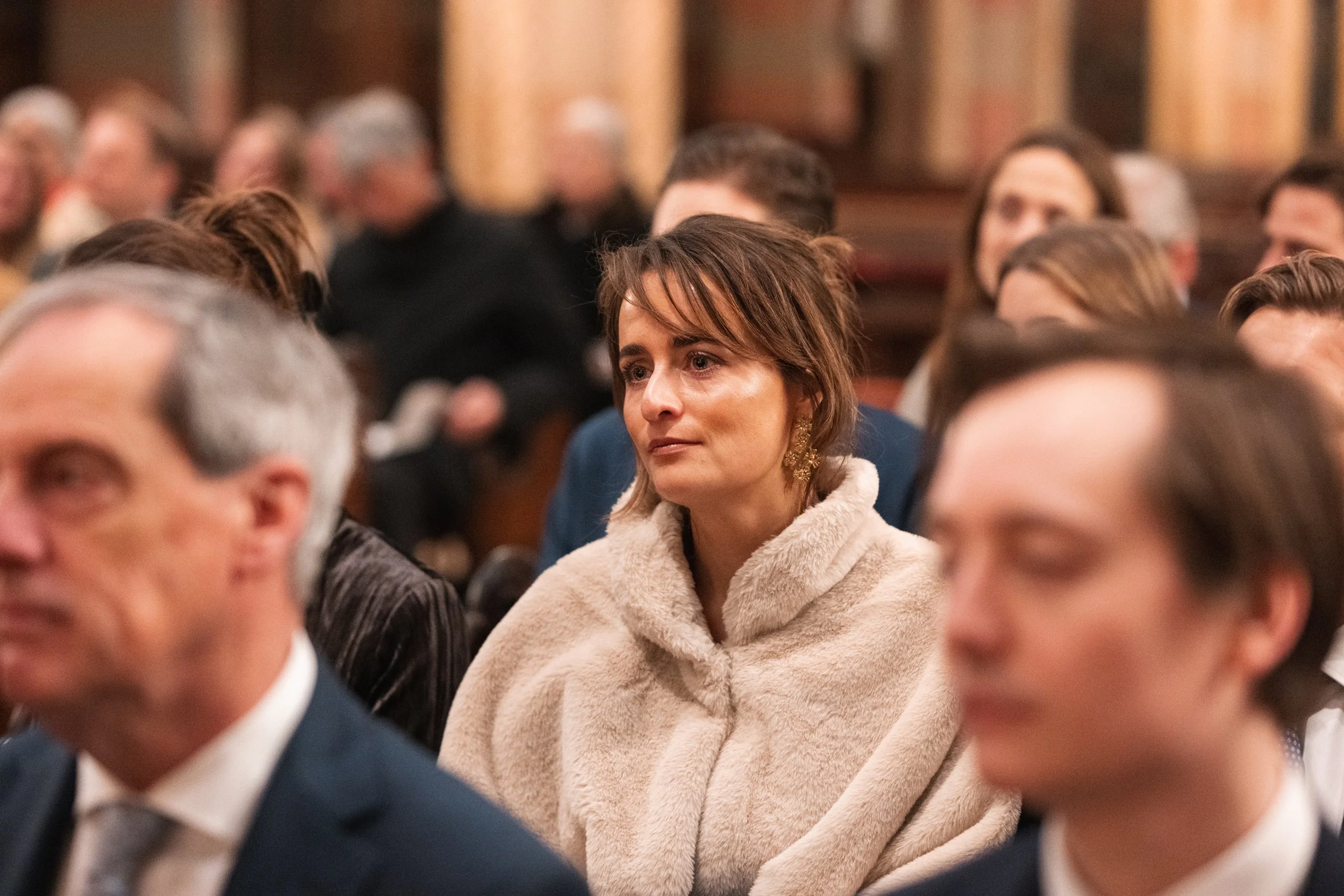 A woman with short brown hair wearing earrings and a beige coat attends an indoor event with many seated people in the background.