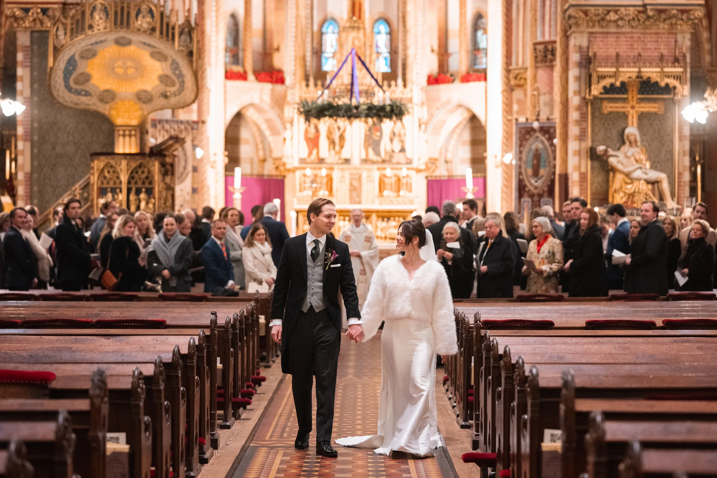 A newlywed couple holding hands walks down the aisle of a church, surrounded by guests. The bride wears a white dress and a fluffy white coat, and the groom is dressed in a formal black suit with a vest and tie. The church interior is decorated with 