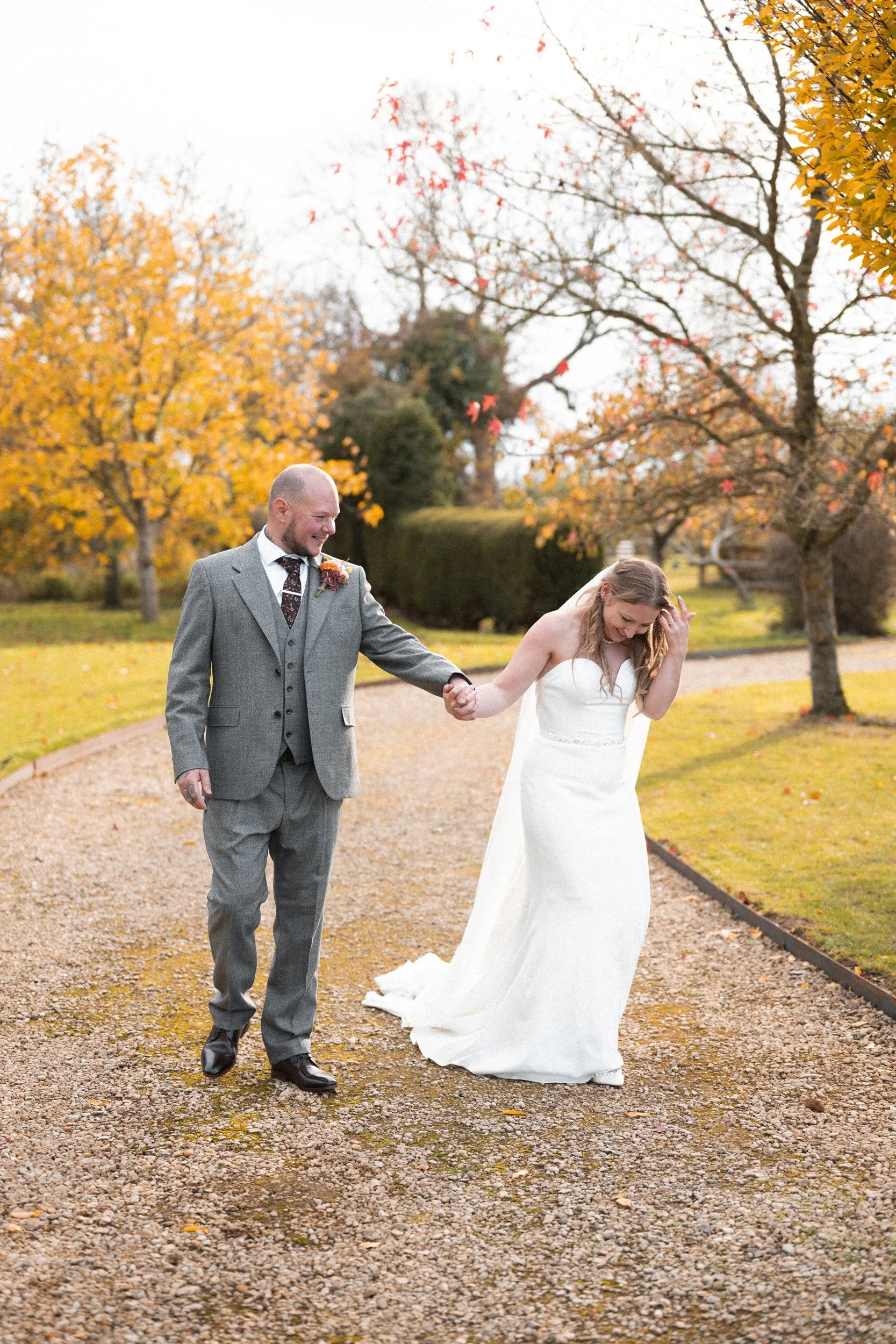 A bride and groom walking hand in hand outdoors in a park with fall foliage, the bride smiling and touching her hair, wearing a strapless white wedding dress, and the groom in a gray suit with a boutonniere.