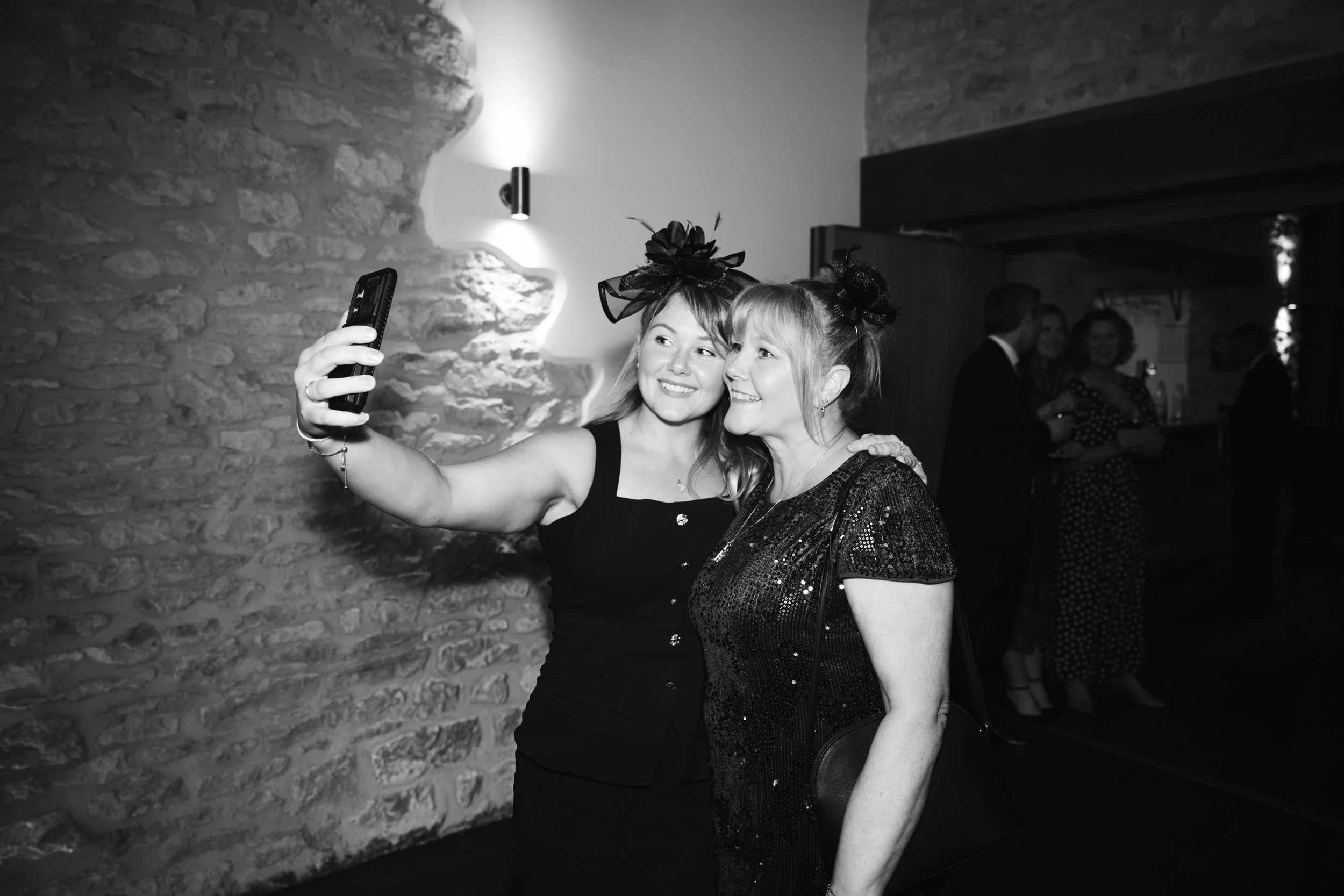 Two women taking a selfie together at a social event, both wearing party dresses and decorative headpieces, in a room with a stone wall and other people in the background.