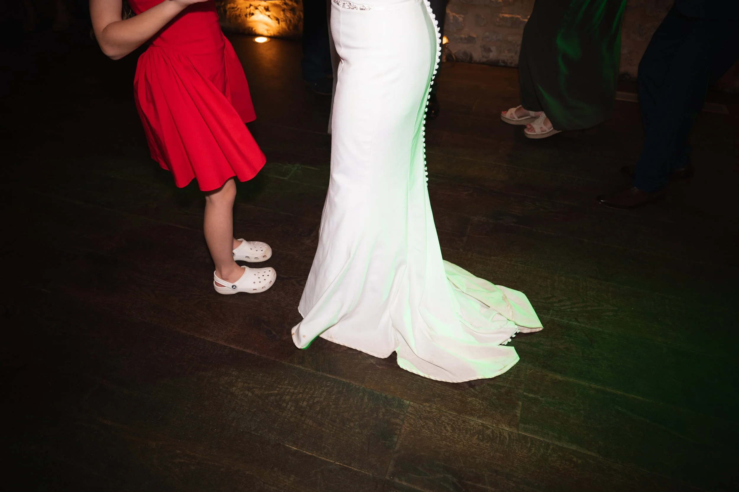 People dancing on a dark wooden floor, including a person in a white wedding dress and a girl in a red dress with white Crocs.