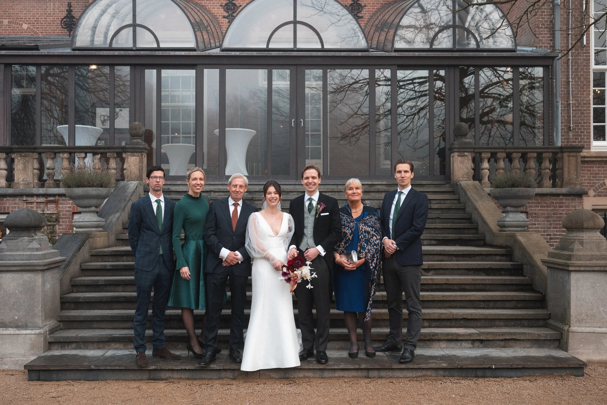 A group of seven people dressed in formal attire, standing on steps outside a brick building with large glass windows and a glass conservatory, during a wedding celebration.