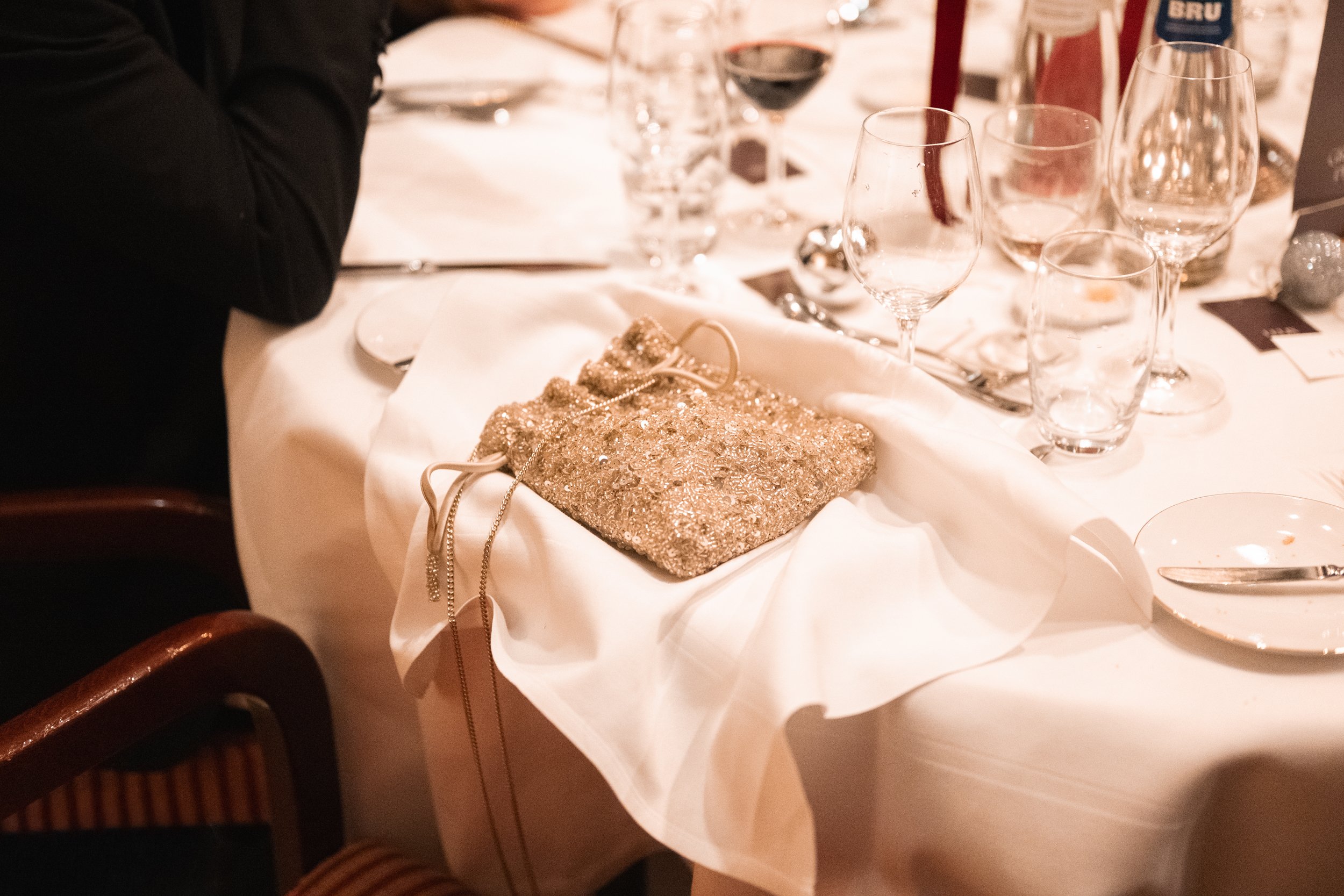 A decorated event table with empty glasses, food plates, and a gold glittery clutch purse with a chain on a white tablecloth.