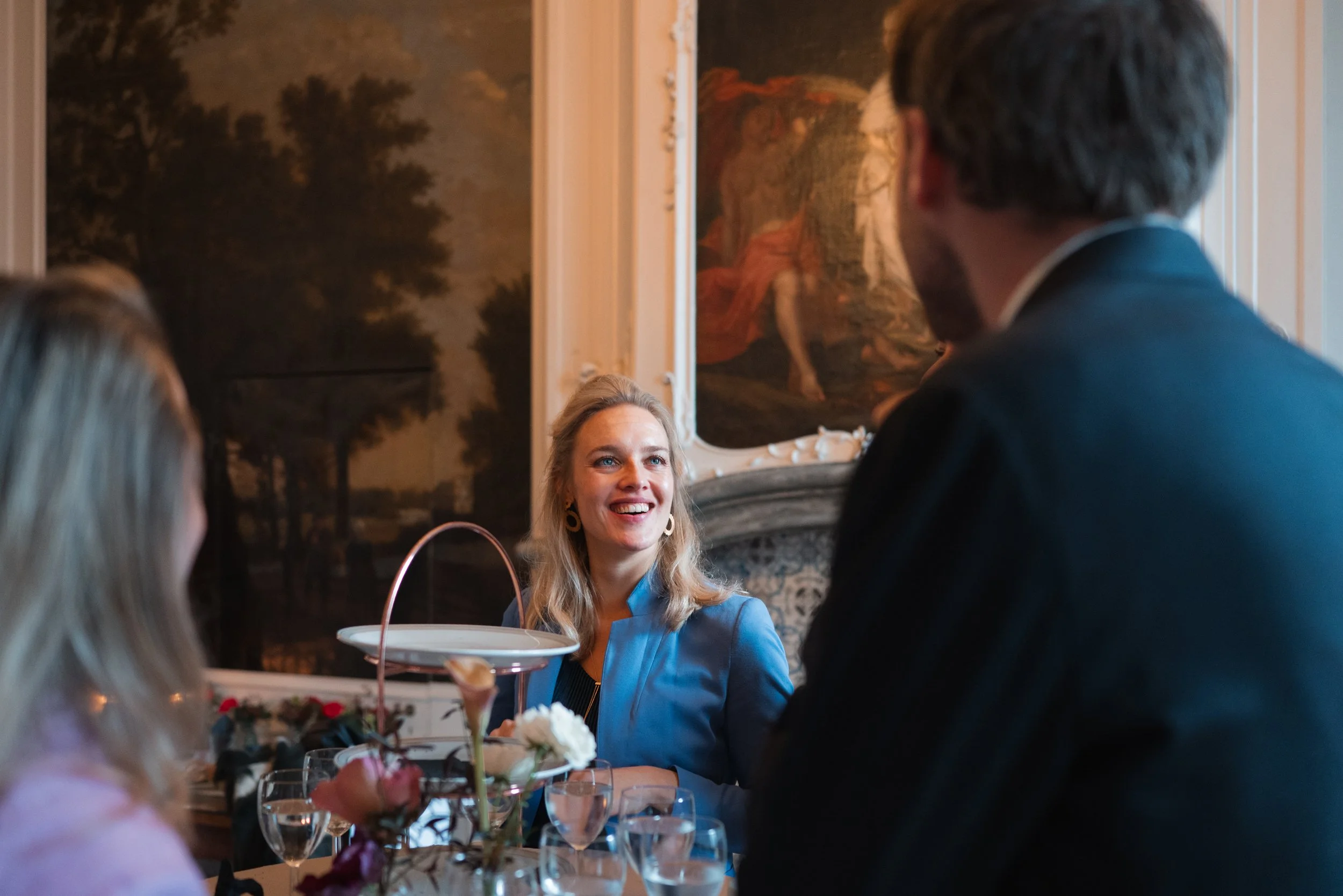 A woman sitting at a dining table with a floral centerpiece, smiling and engaging in conversation with a man and a girl at a formal gathering or dinner event in a room decorated with classical paintings.