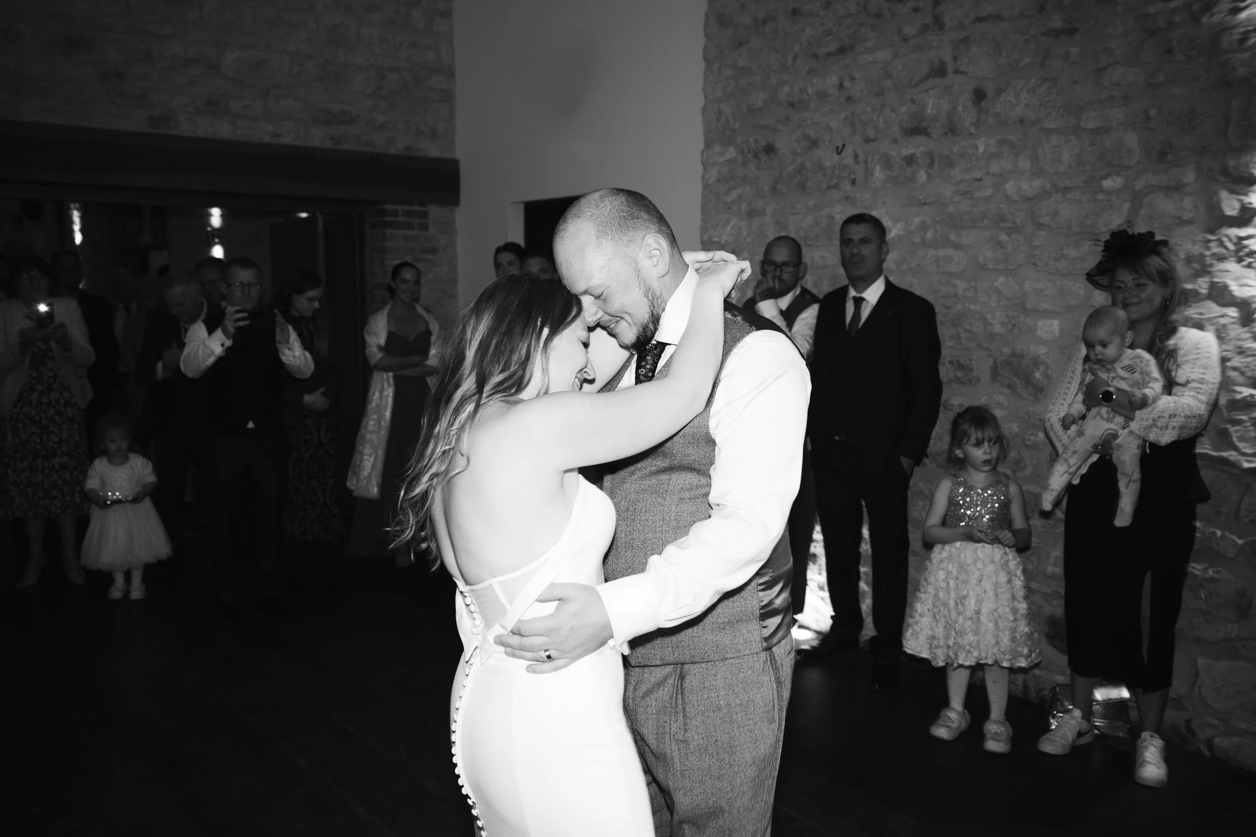 Black and white photo of a bride and groom dancing closely, with wedding guests watching in the background.