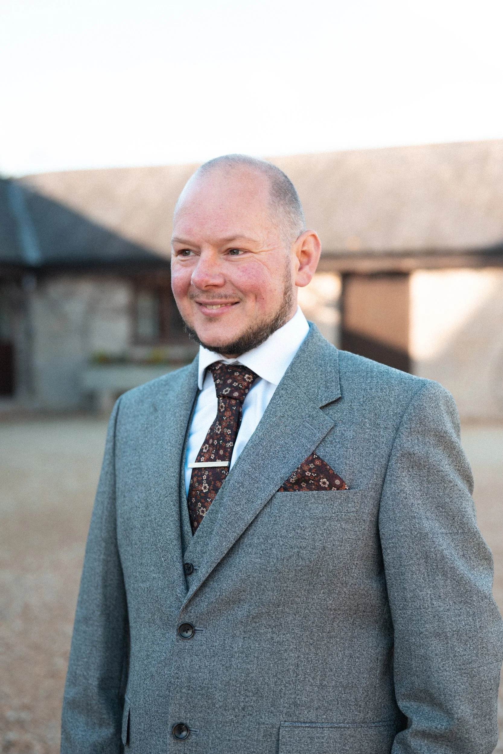 A man in a gray suit, white shirt, and patterned tie with a matching pocket square, smiling outdoors during daylight with a building in the background.