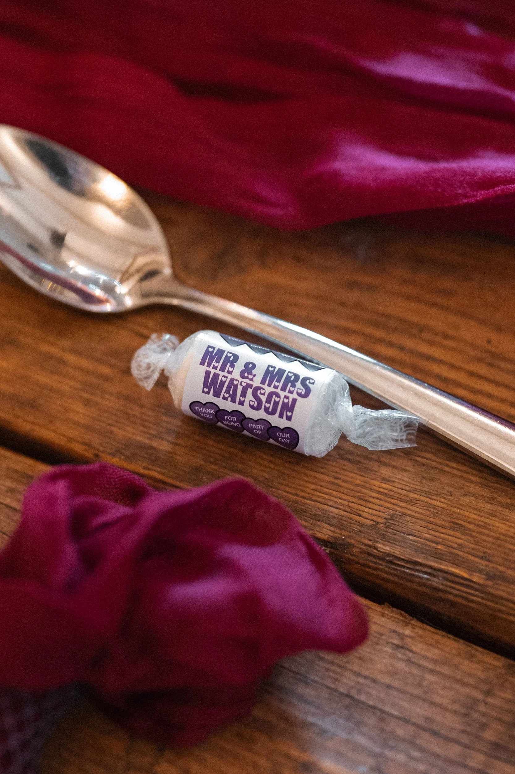 A candy wrapped with a label reading 'Mr. & Mrs. Watson' resting on a wooden table next to a silver spoon and a burgundy cloth.