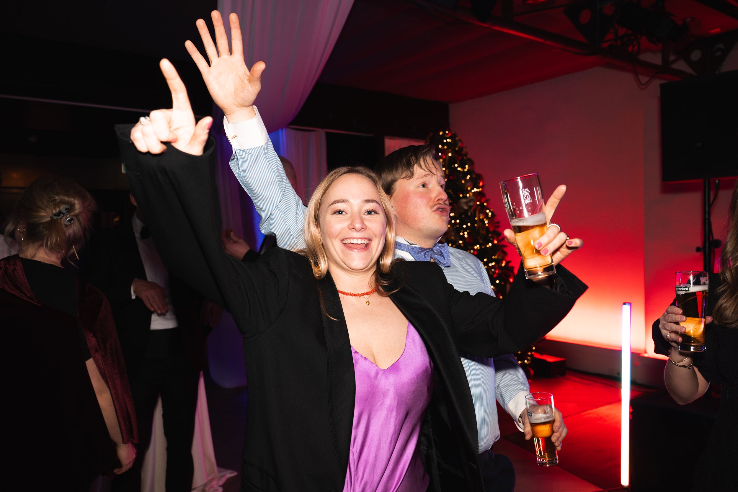 People celebrating at a party with a Christmas tree in the background, holding drinks and smiling.