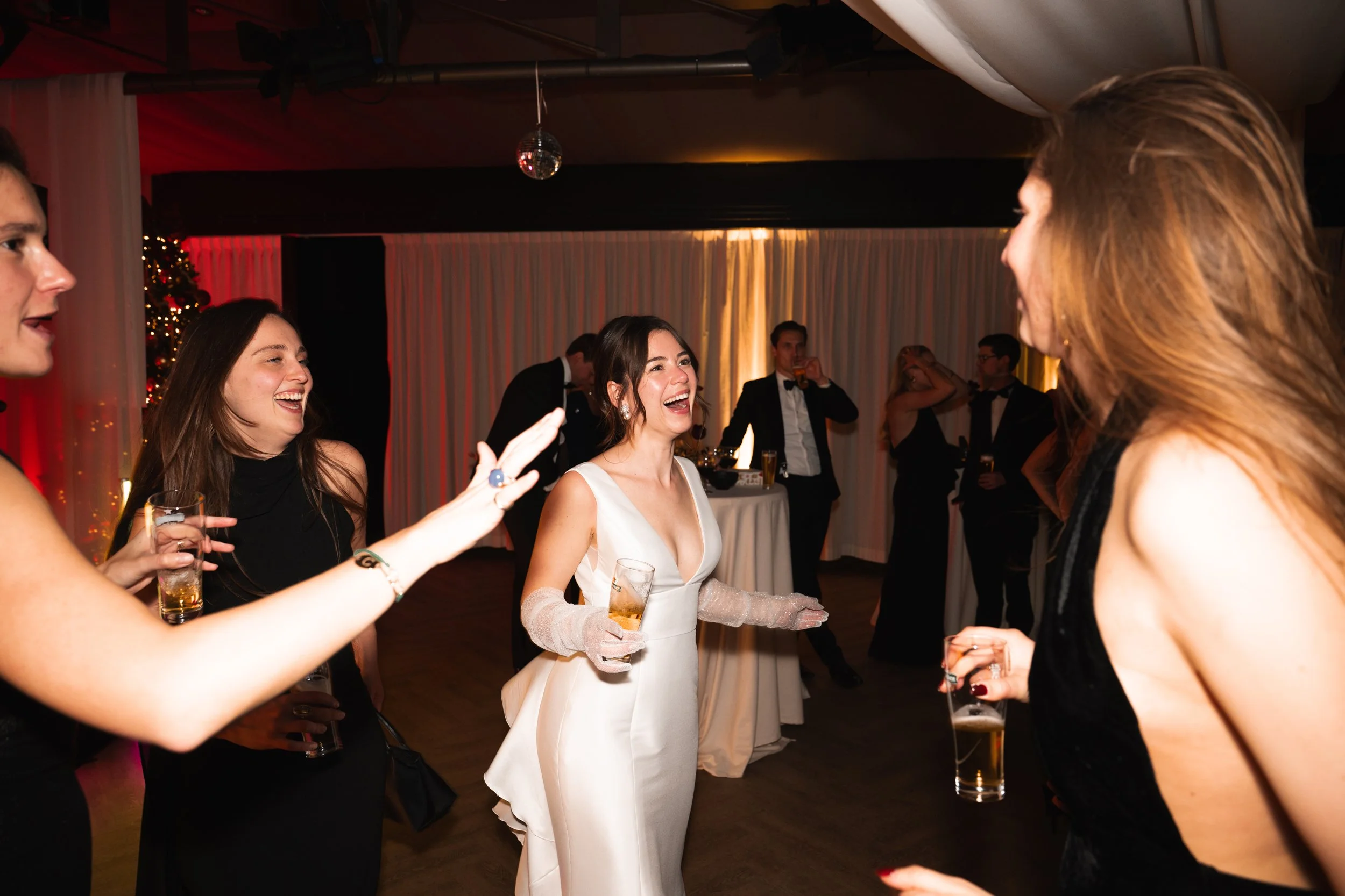 People dancing and celebrating at a wedding reception, with a bride in a white dress laughing and holding a drink, surrounded by friends in formal attire.