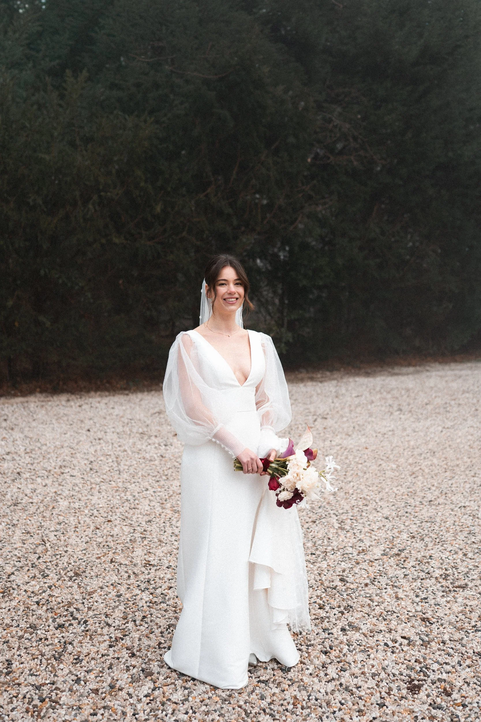 A bride in a white wedding dress holding a bouquet of flowers, standing outdoors on a gravel surface with trees in the background.