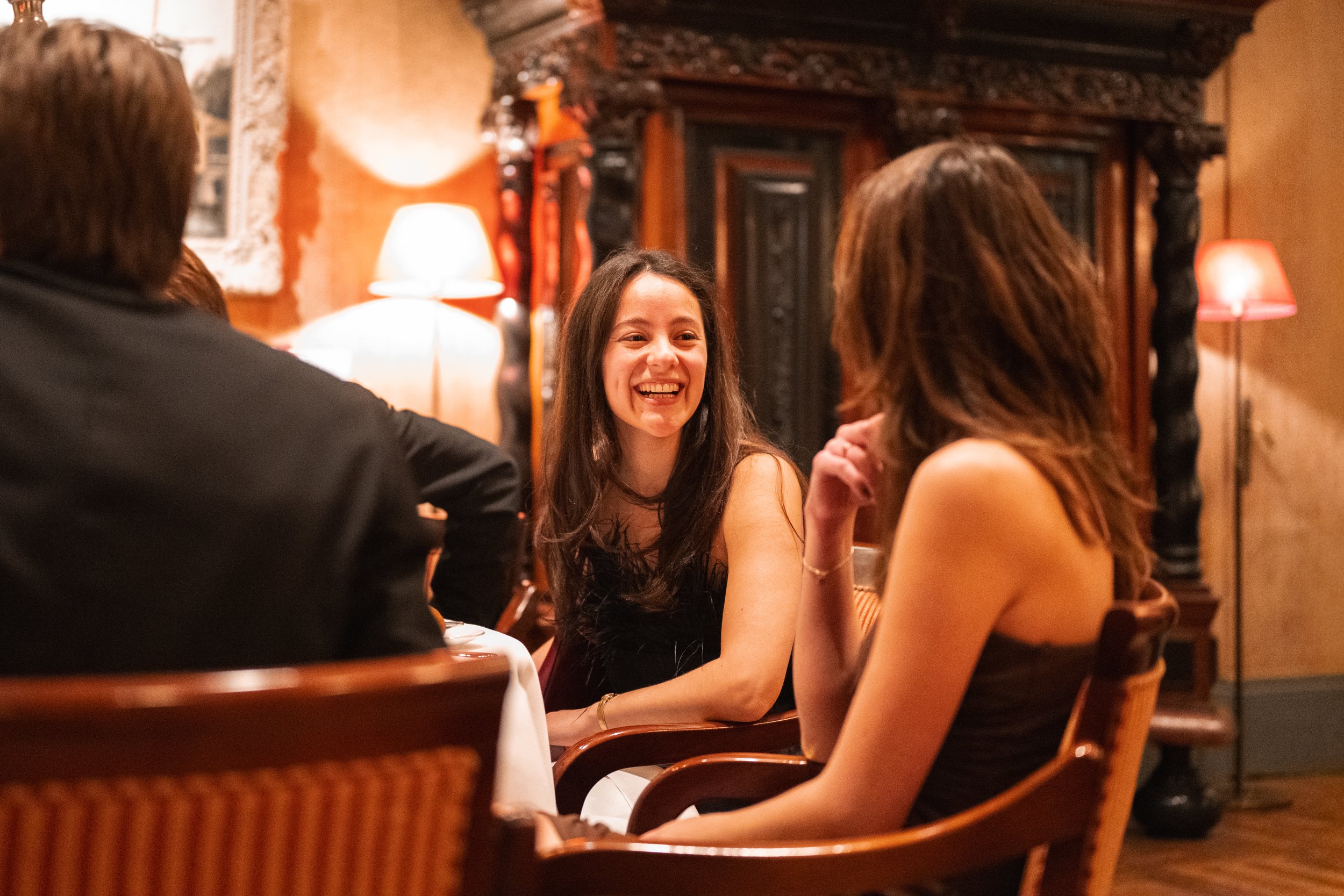 A group of people sitting at a table in a warmly lit restaurant, engaged in conversation and smiling.