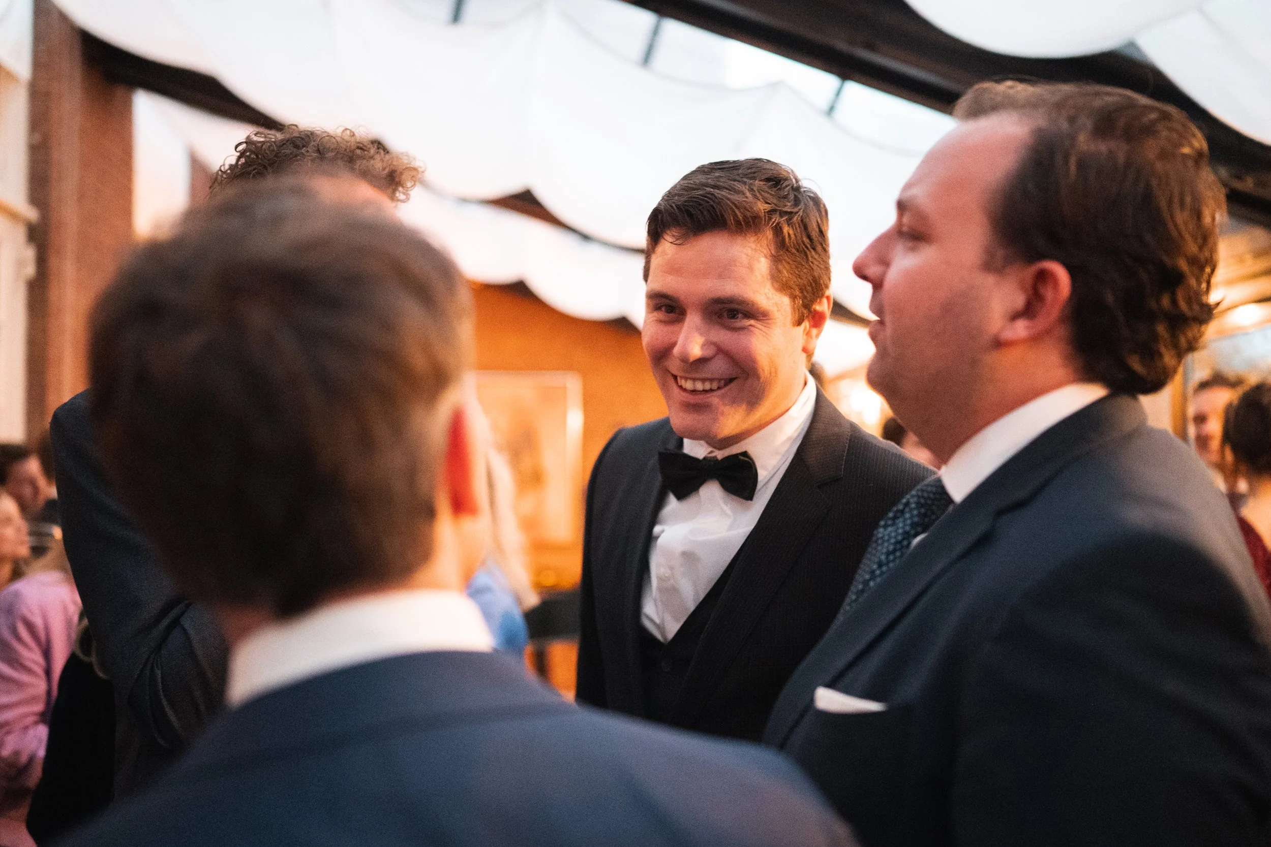 Four men in tuxedos engaged in a conversation at a formal event with a decorated indoor setting.