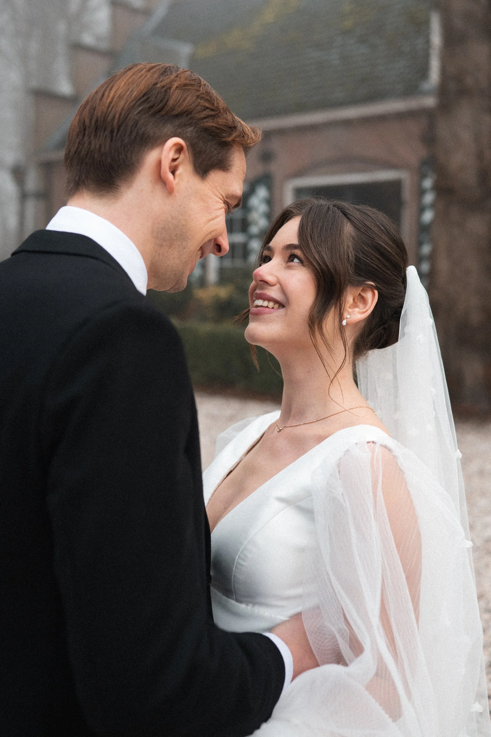 A couple on their wedding day, smiling and looking into each other's eyes outdoors.