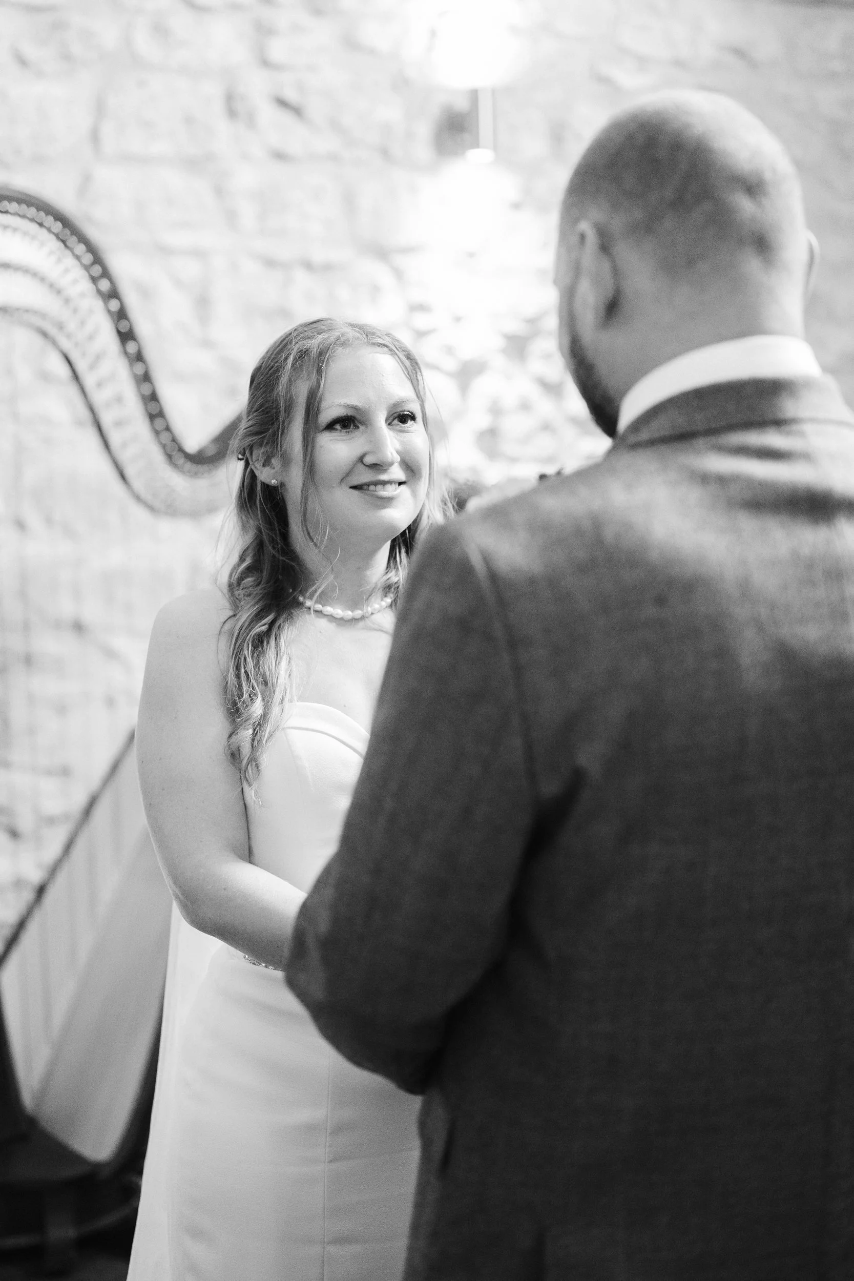 A woman in a wedding dress and pearl necklace smiling at a man in a suit during a wedding ceremony.