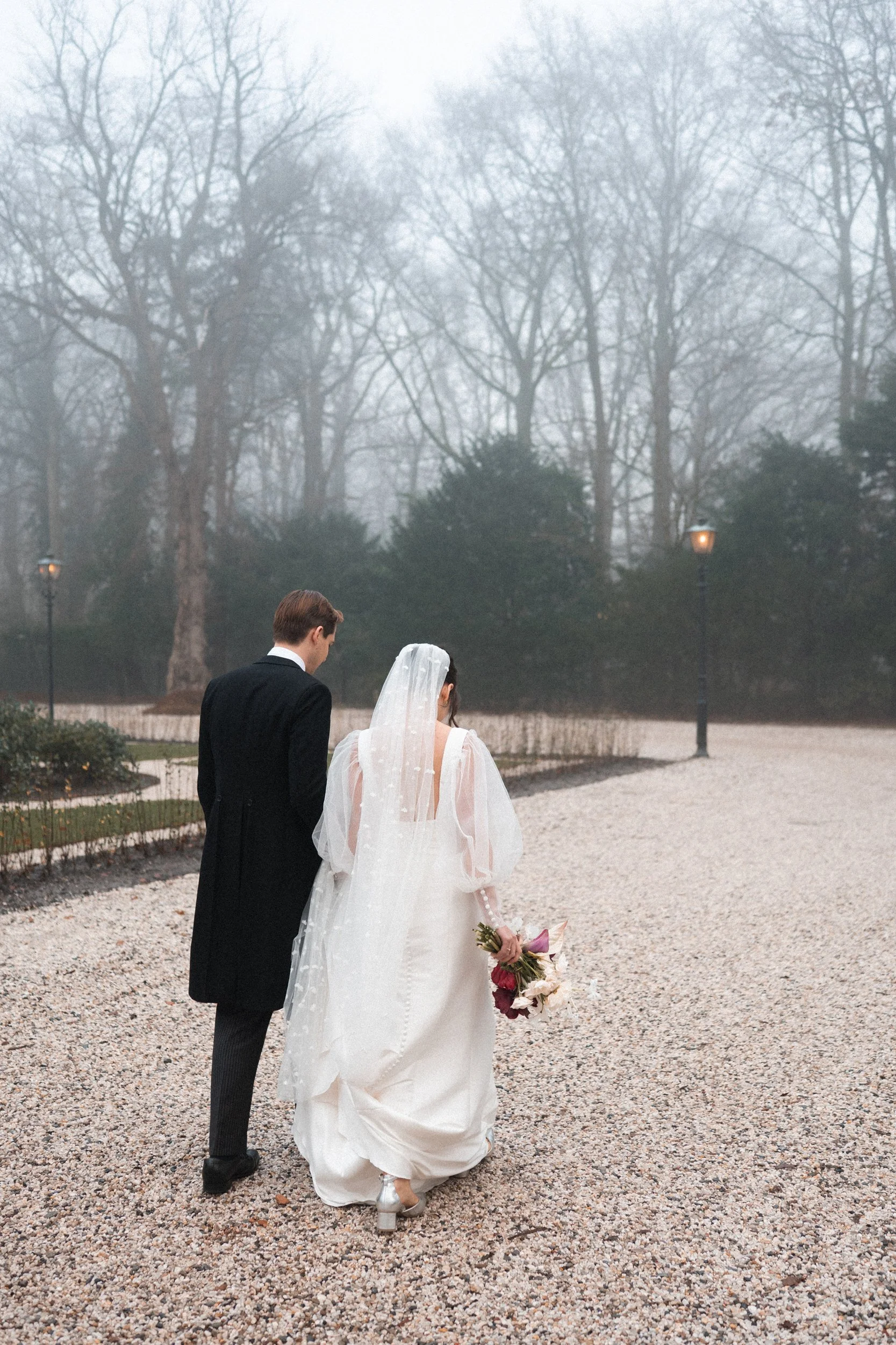 A bride and groom walking together outdoors on a foggy day, with the bride holding a bouquet of flowers and wearing a white wedding dress, and the groom dressed in a black suit.