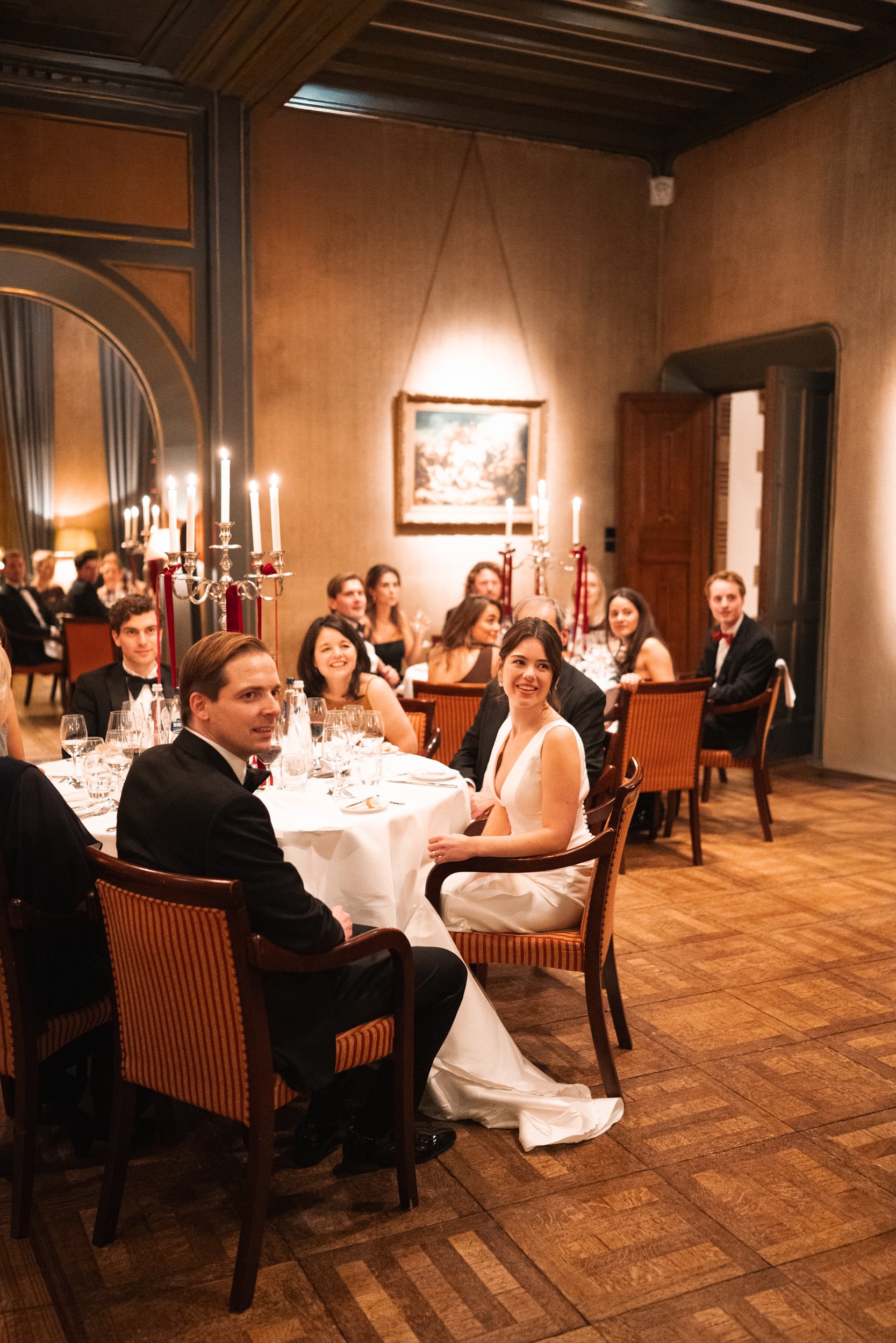 Guests in formal attire seated at a wedding reception table in an elegant, warmly lit room with candelabras and artwork