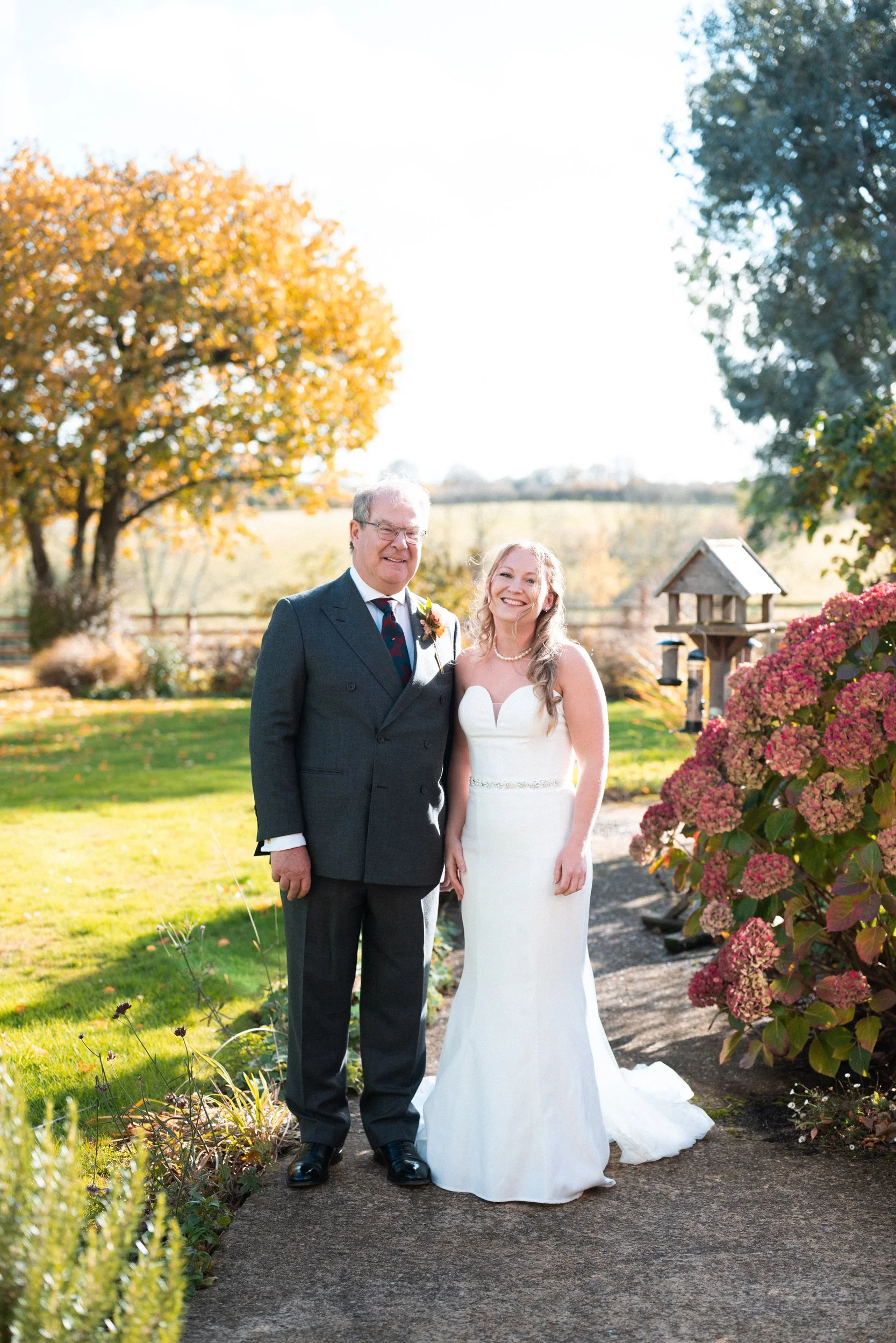 A bride in a white wedding gown standing next to an older man in a suit outdoors on a sunny day, with fall foliage and pink hydrangeas in the background.