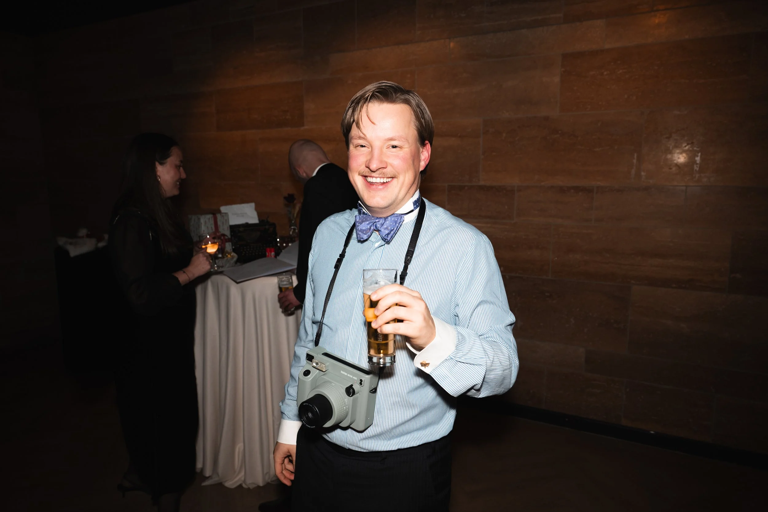 A man in a blue shirt with a camera around his neck, holding a drink, smiling at a party with two people in the background near a table.