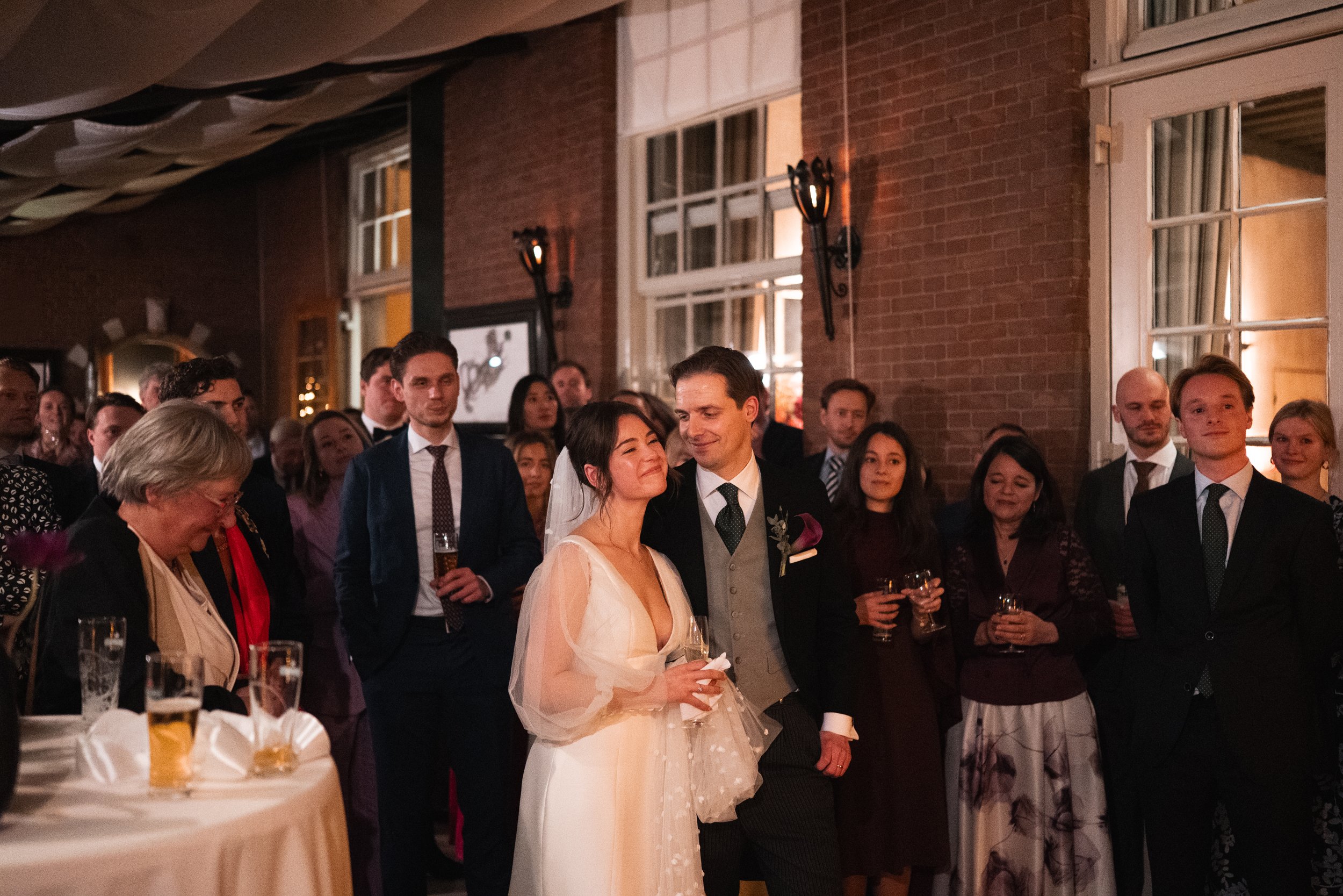 A wedding celebration with a bride and groom in the center, surrounded by guests in formal attire. The bride is smiling and holding a glass, while the groom leans towards her. The setting appears to be indoors in a decorated venue with warm lighting.