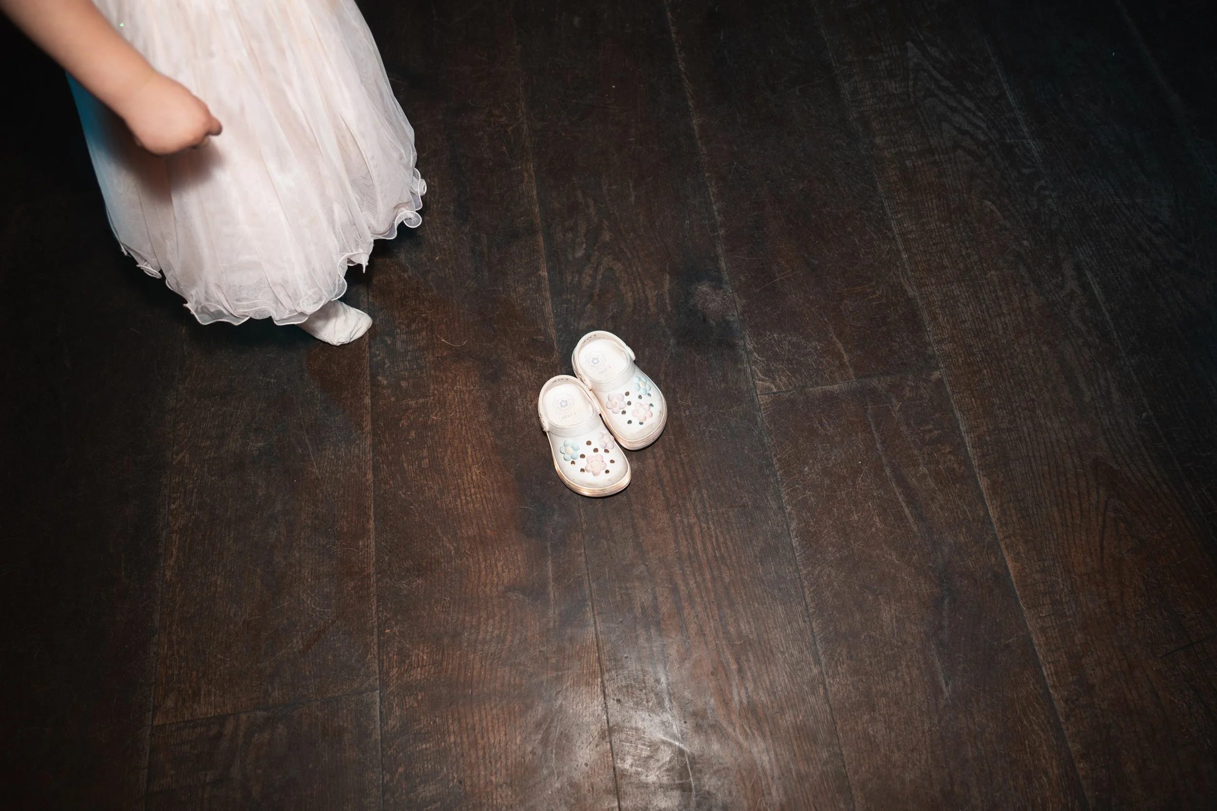 A pair of children's white slip-on shoes with floral patterns on a dark wooden floor, with part of a child's white dress and foot visible on the left side of the image.