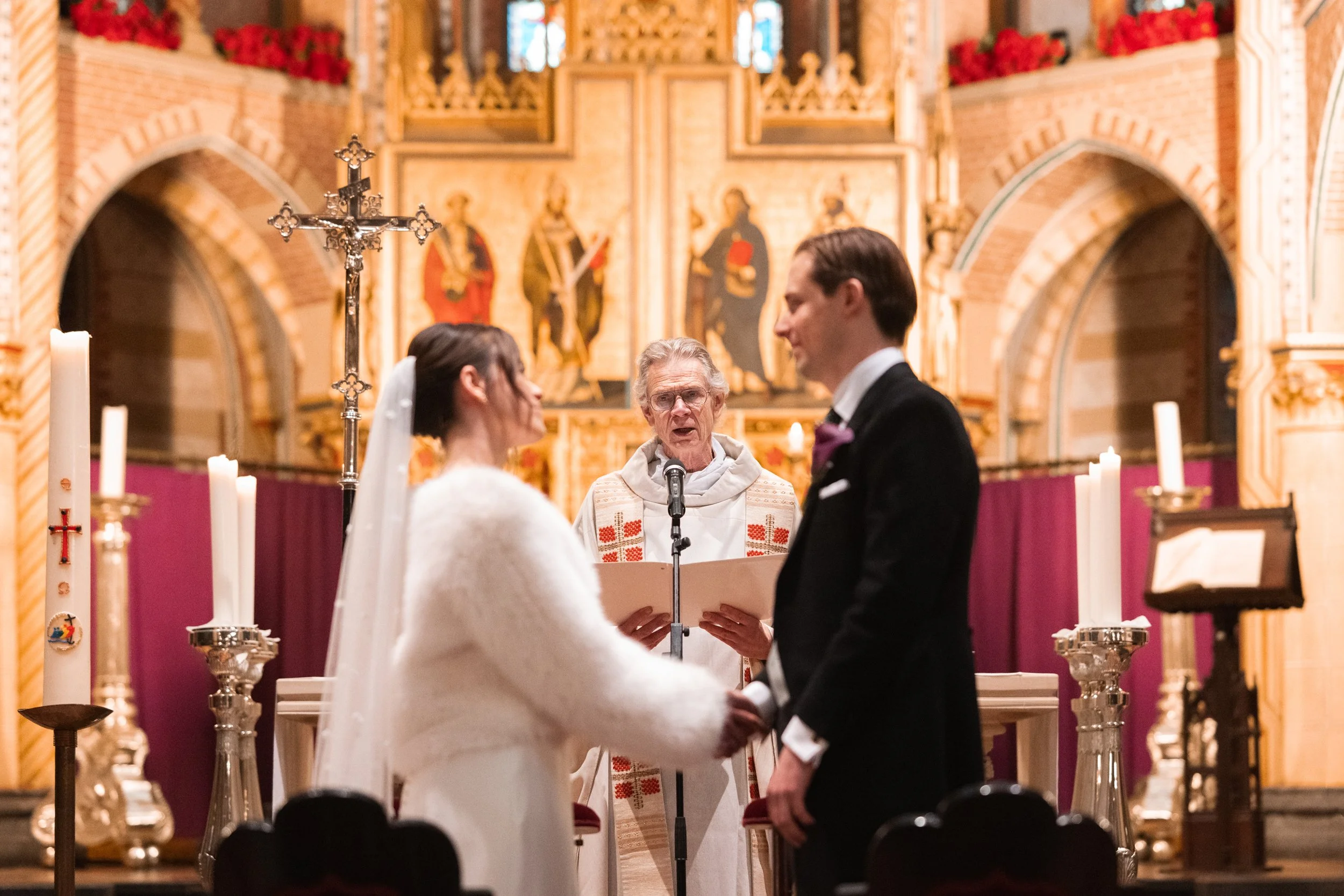 A bride and groom holding hands at their wedding ceremony in a church, with a priest officiating in the background. The church has ornate decorations, candles, and religious artwork.