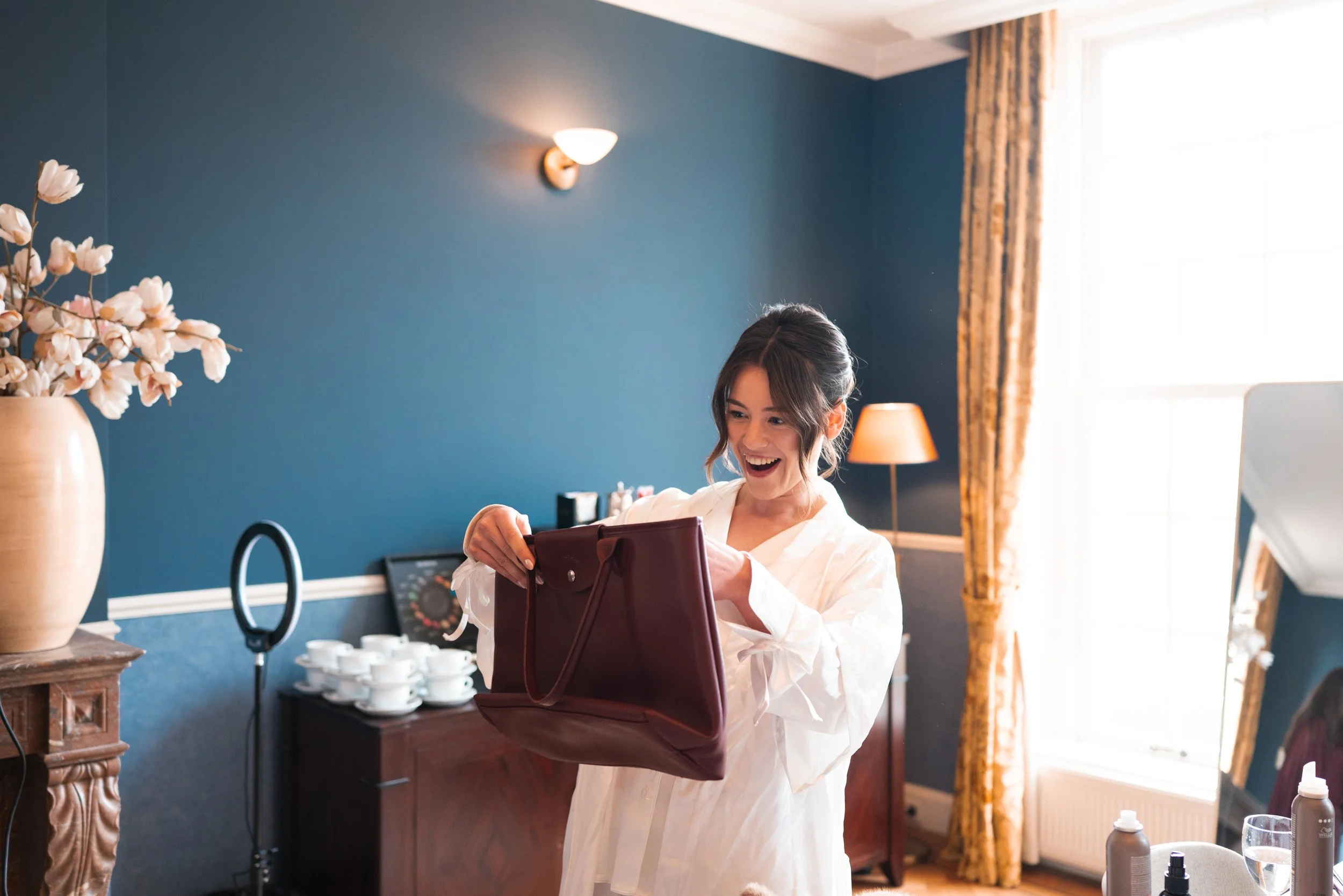 A woman with dark hair in a loose bun, wearing a white robe, smiles and looks inside a burgundy handbag in a well-lit room with a blue wall, a vase of pink flowers, a wooden sideboard with white ceramics, and a large window with yellow curtains.