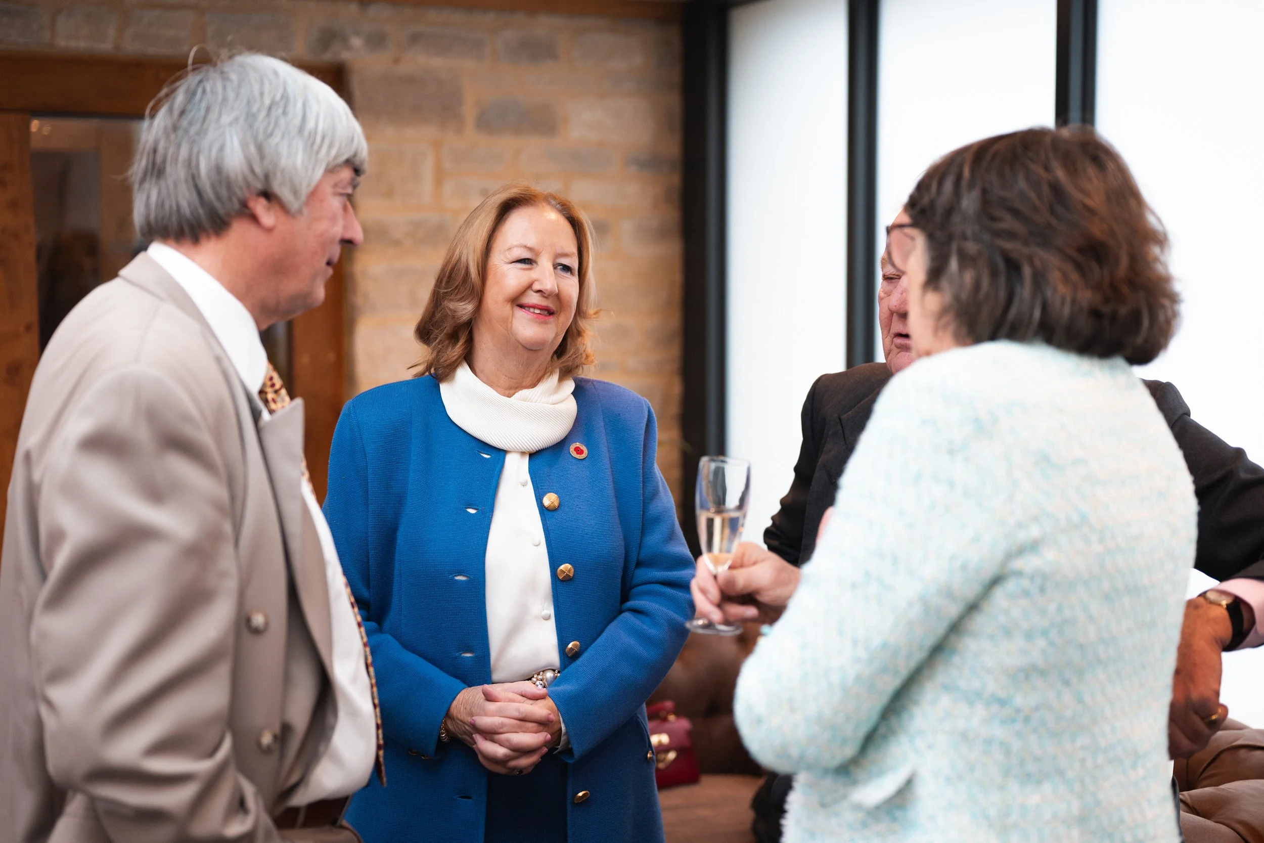 A group of four older adults engaging in conversation at a social gathering, with one woman holding a glass of champagne, indoors with a brick wall and windows in the background.