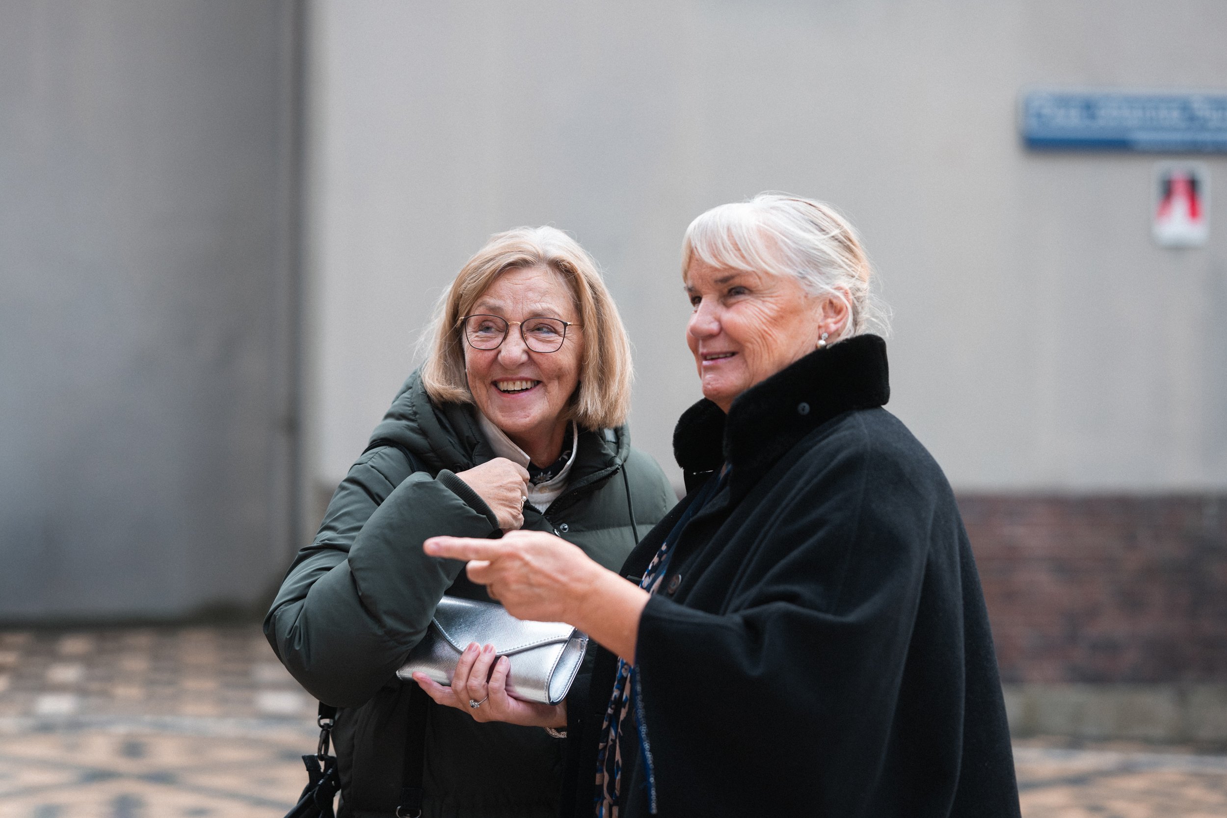 Two older women smiling and talking outside. One woman is wearing glasses and a green jacket, holding a silver purse. The other woman has white hair, a black coat, and is pointing while smiling.