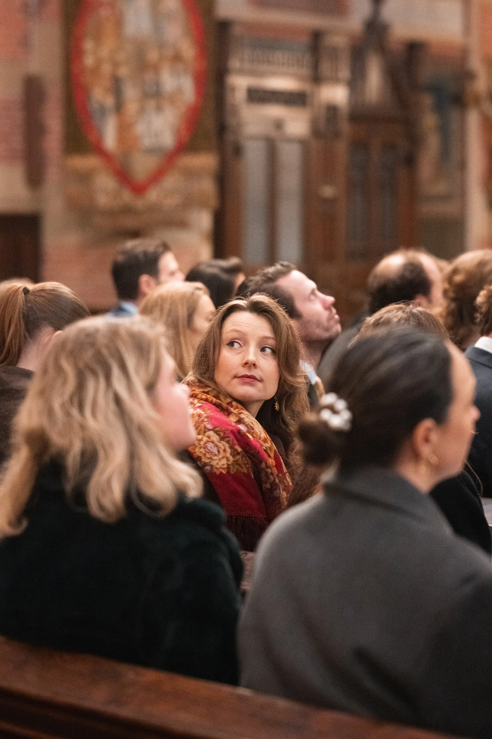 A woman with curly brown hair and a floral scarf looking over her shoulder in a crowded indoor setting, possibly a lecture or church.