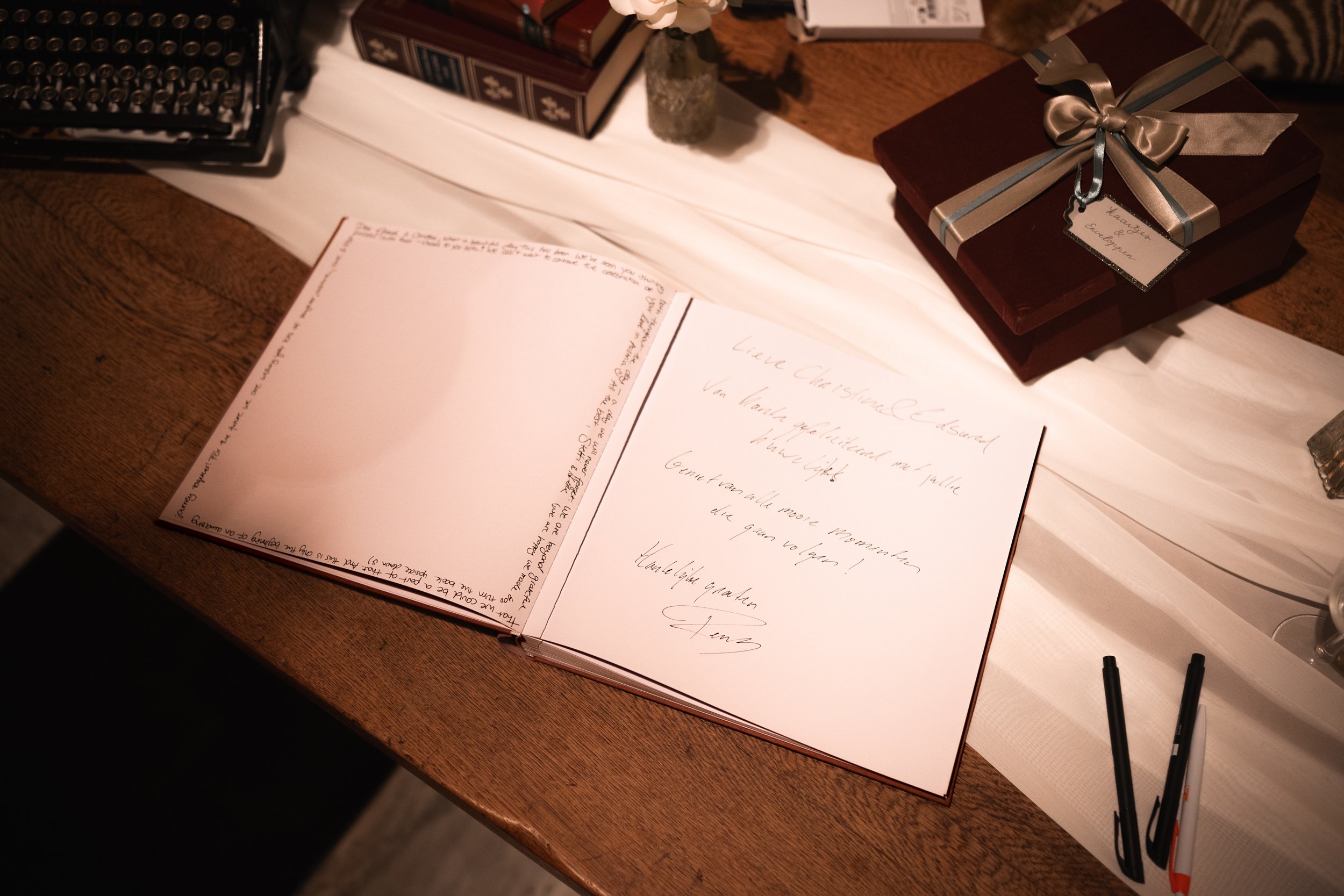 A wooden table with a white cloth cover, holding a guestbook with handwritten messages, a closed gift box wrapped with a silver ribbon, two black pens, and other decorative items in the background.