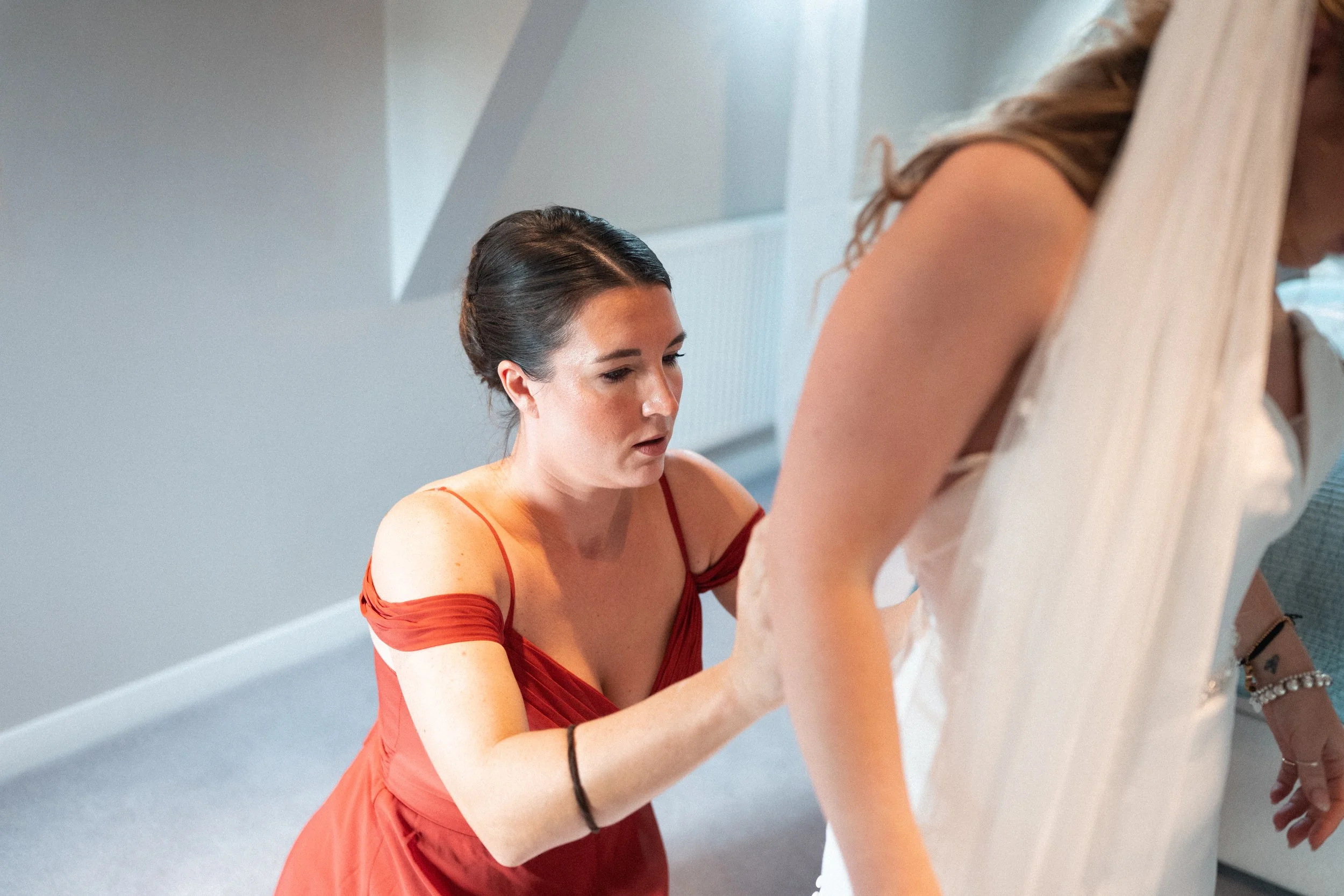 A woman in a red dress assists another woman in a white dress with her outfit, possibly during preparations for a wedding.