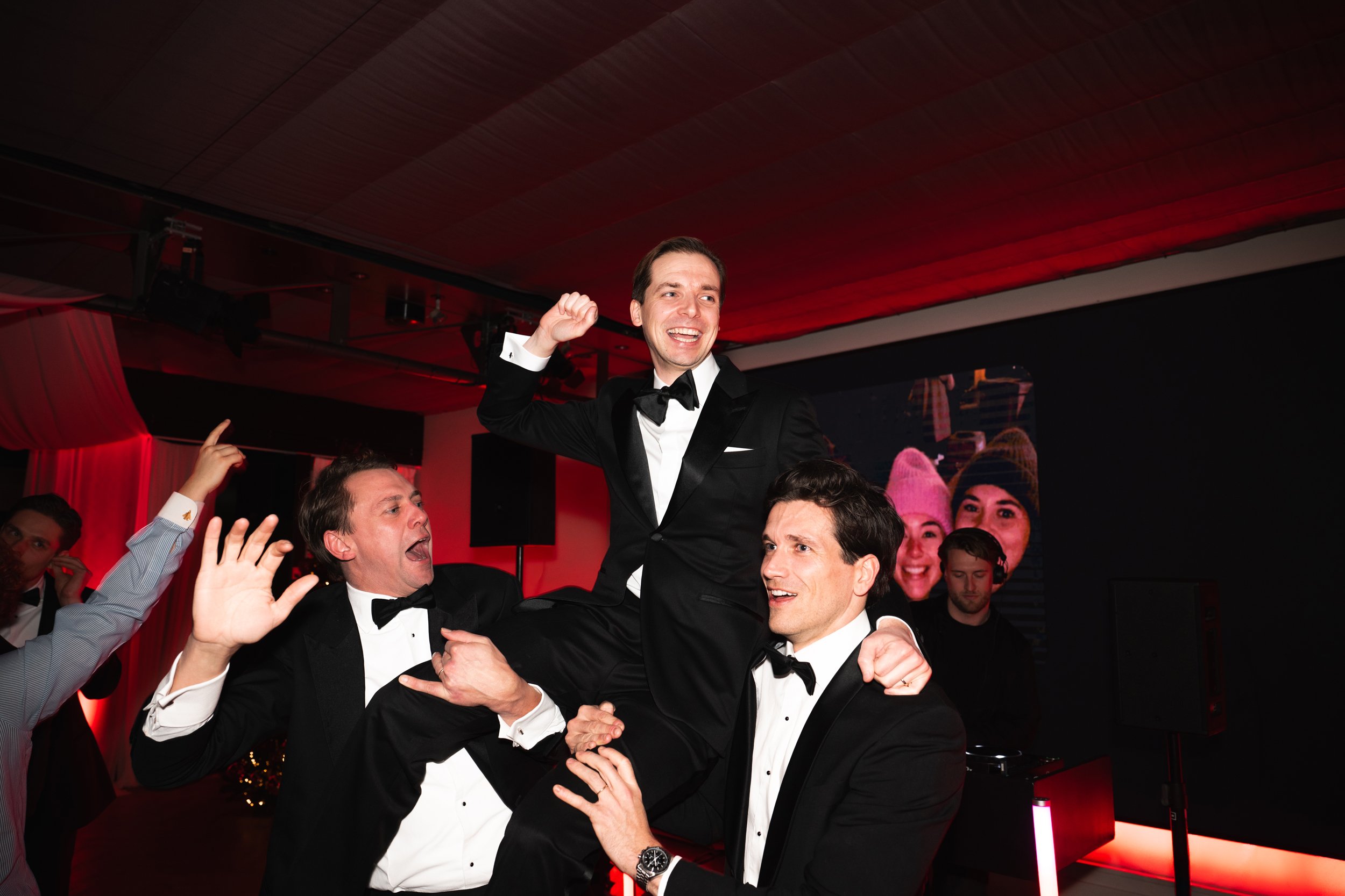 Men in tuxedos celebrating at a formal event, lifting a smiling man in a tuxedo and bowtie in the air. Others around are cheering and dancing.
