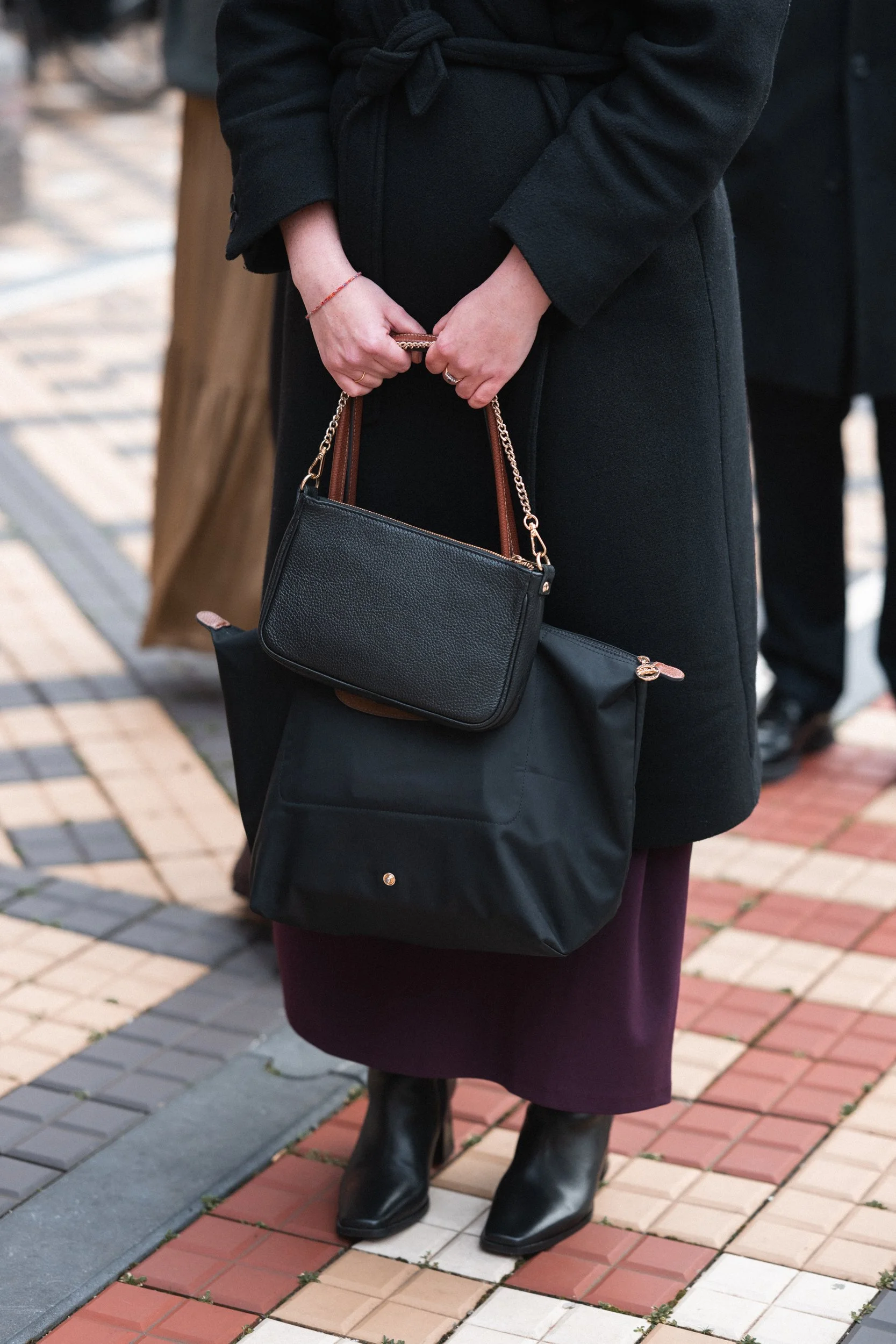 Person wearing black coat and boots holding a black purse and a black bag, standing on a sidewalk with a tiled pattern.