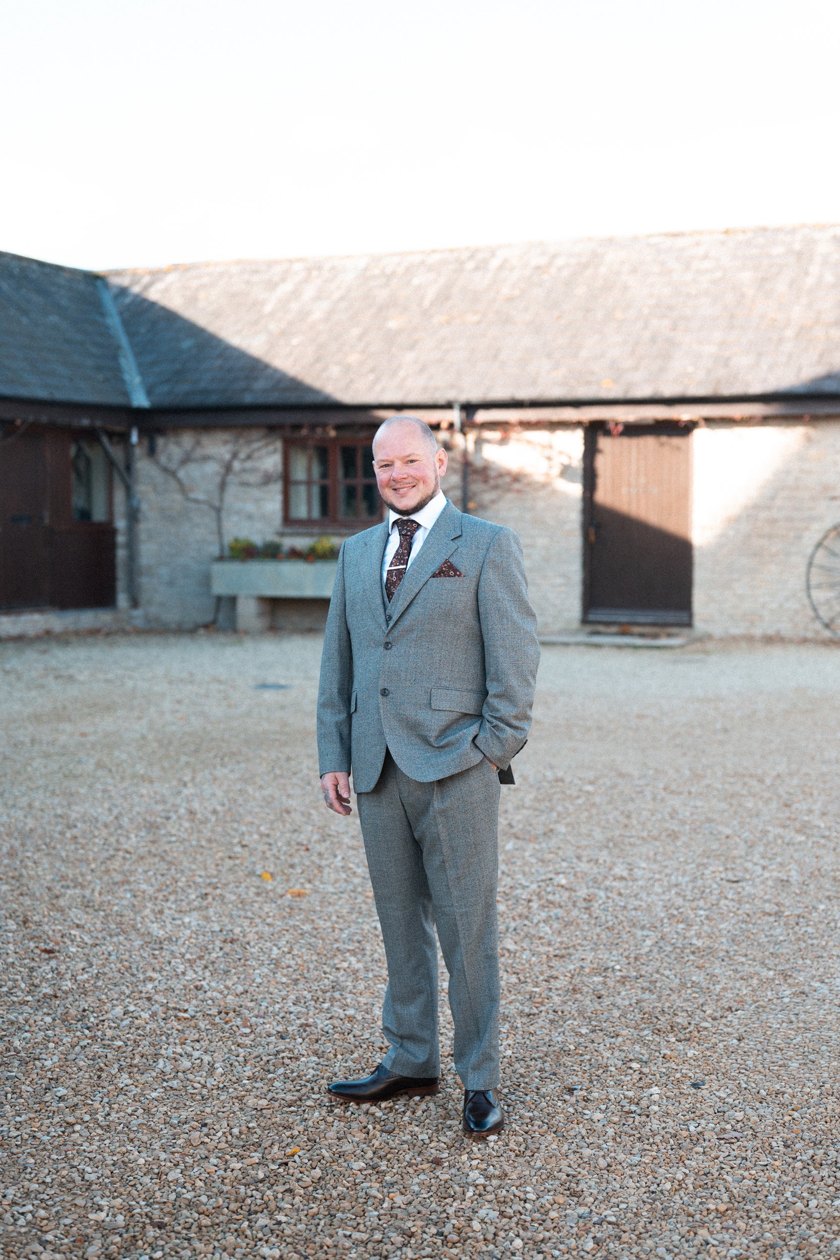 A man in a gray suit standing outdoors on a gravel surface, smiling, with a rustic building and clear sky in the background.