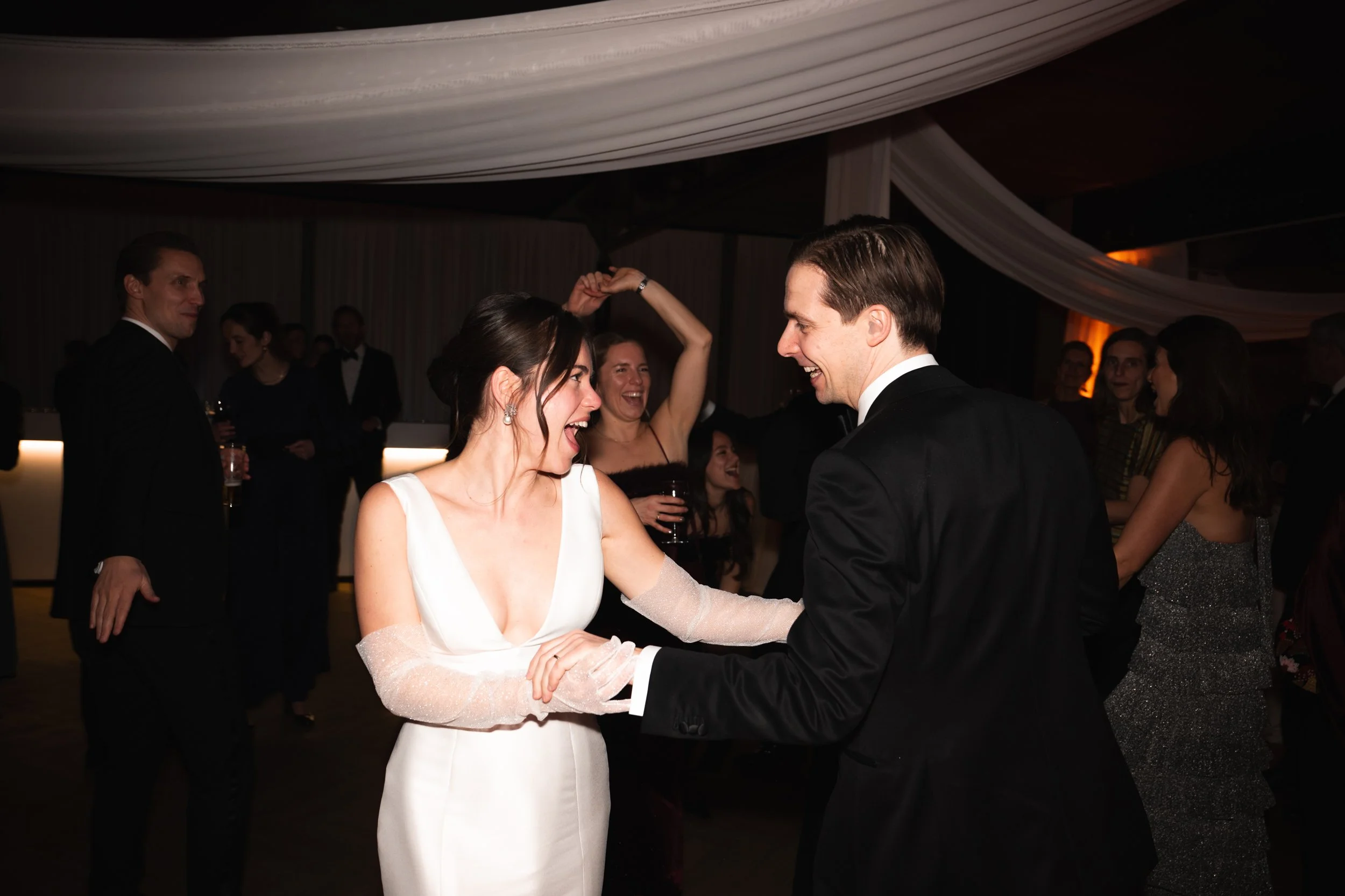 A bride and groom dancing and smiling at their wedding reception, surrounded by guests in formal attire.