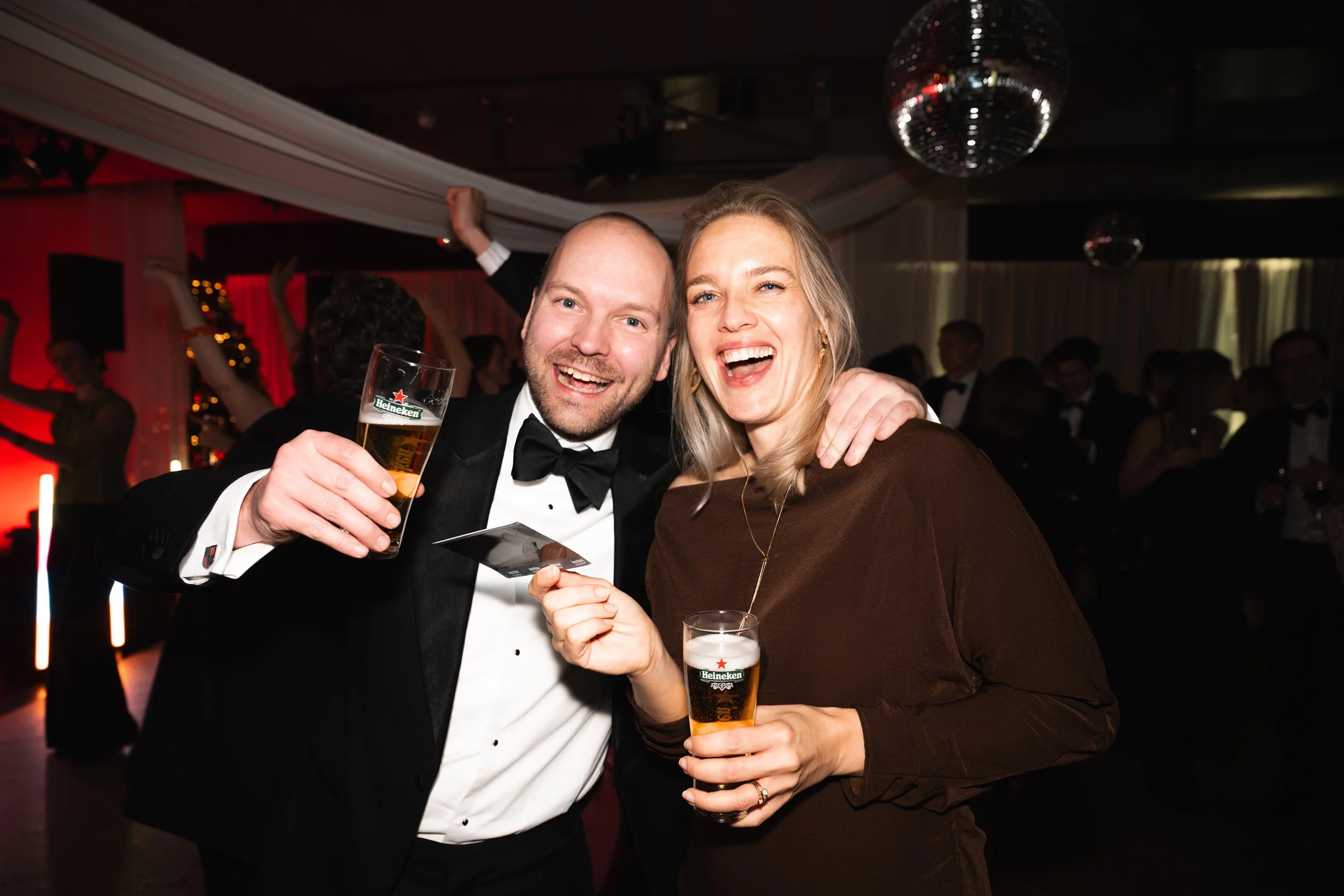 A man and woman dressed in formal attire celebrating at a party, holding drinks and smiling.