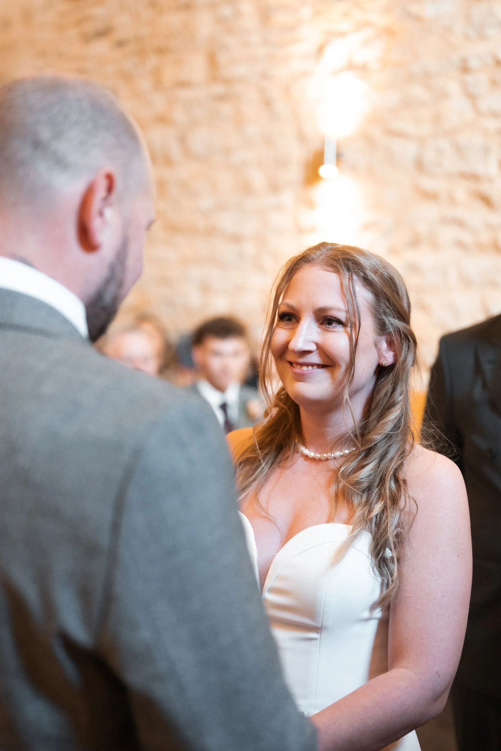 A woman with long wavy hair wearing a white dress and pearl necklace smiles at a man with a beard and in a suit, during a wedding ceremony in a warmly lit room with a brick wall background.