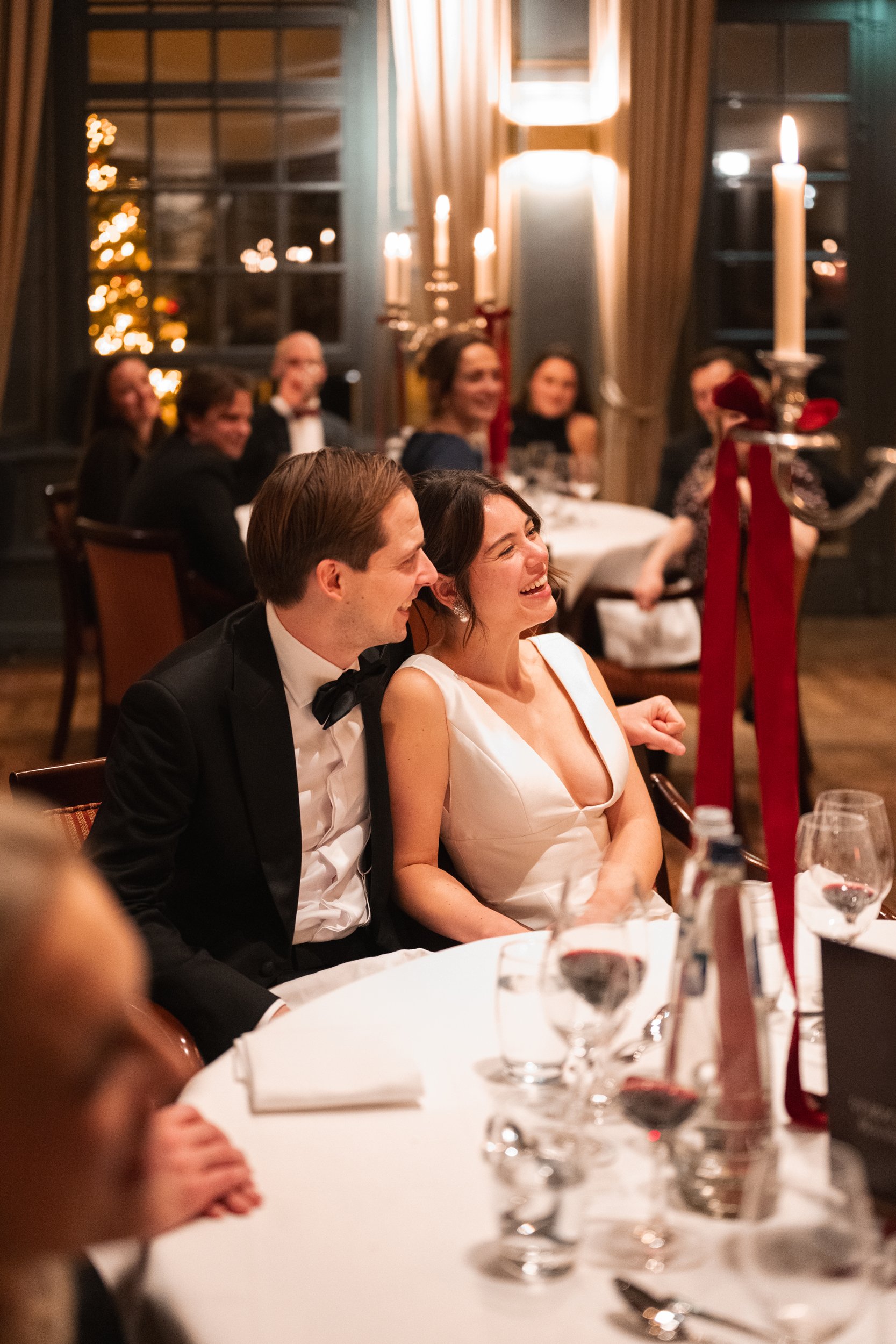 A joyful couple, a man in a black tuxedo and a woman in a white dress, sitting close together at a formal dinner party, smiling and laughing, surrounded by friends and family in an elegant decorated room with candles and a Christmas tree in the backg